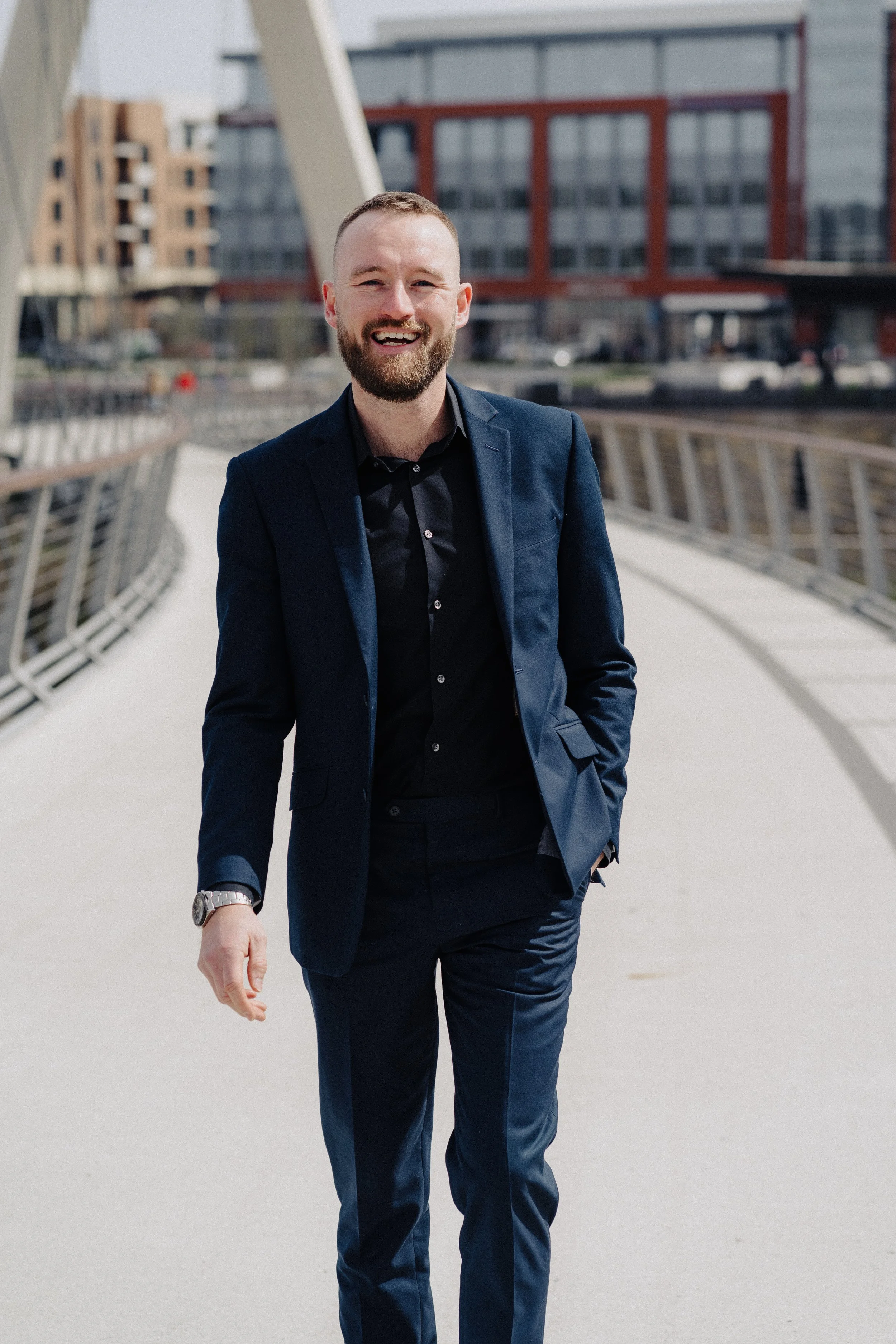 A man in a navy blue suit walking outdoors on a modern bridge with city buildings in the background, smiling.