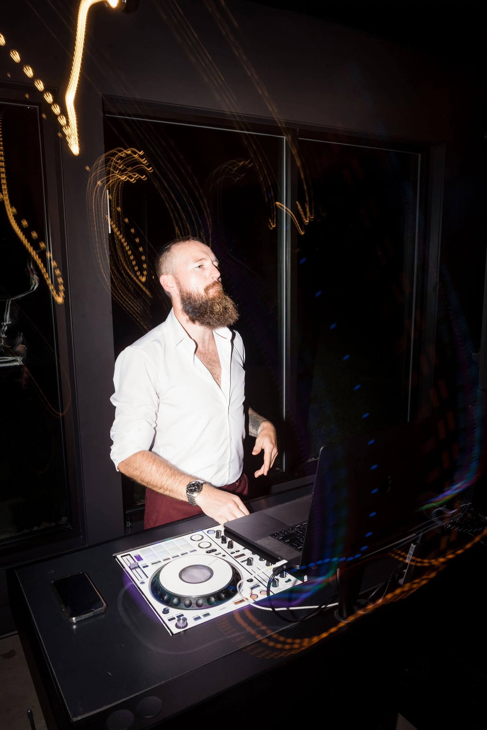 A man with a beard, wearing a white shirt and maroon pants, standing behind DJ equipment in a dark room with reflective glass panels. Light streaks are visible due to long exposure.