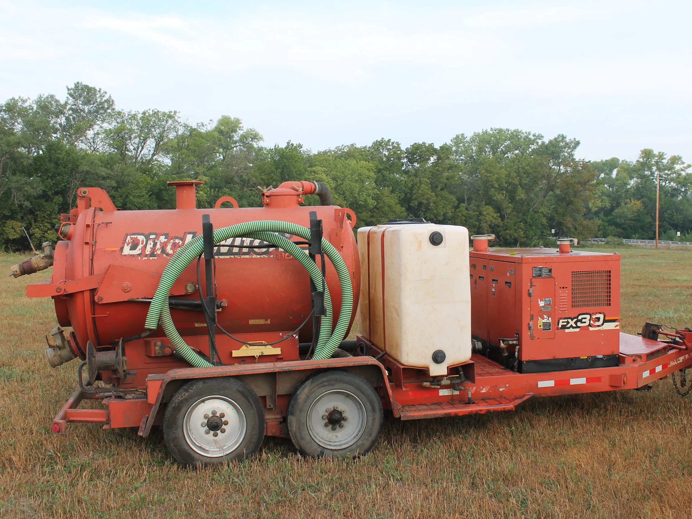 Red industrial equipment on a trailer in a grassy field with trees in the background and a cloudy sky.