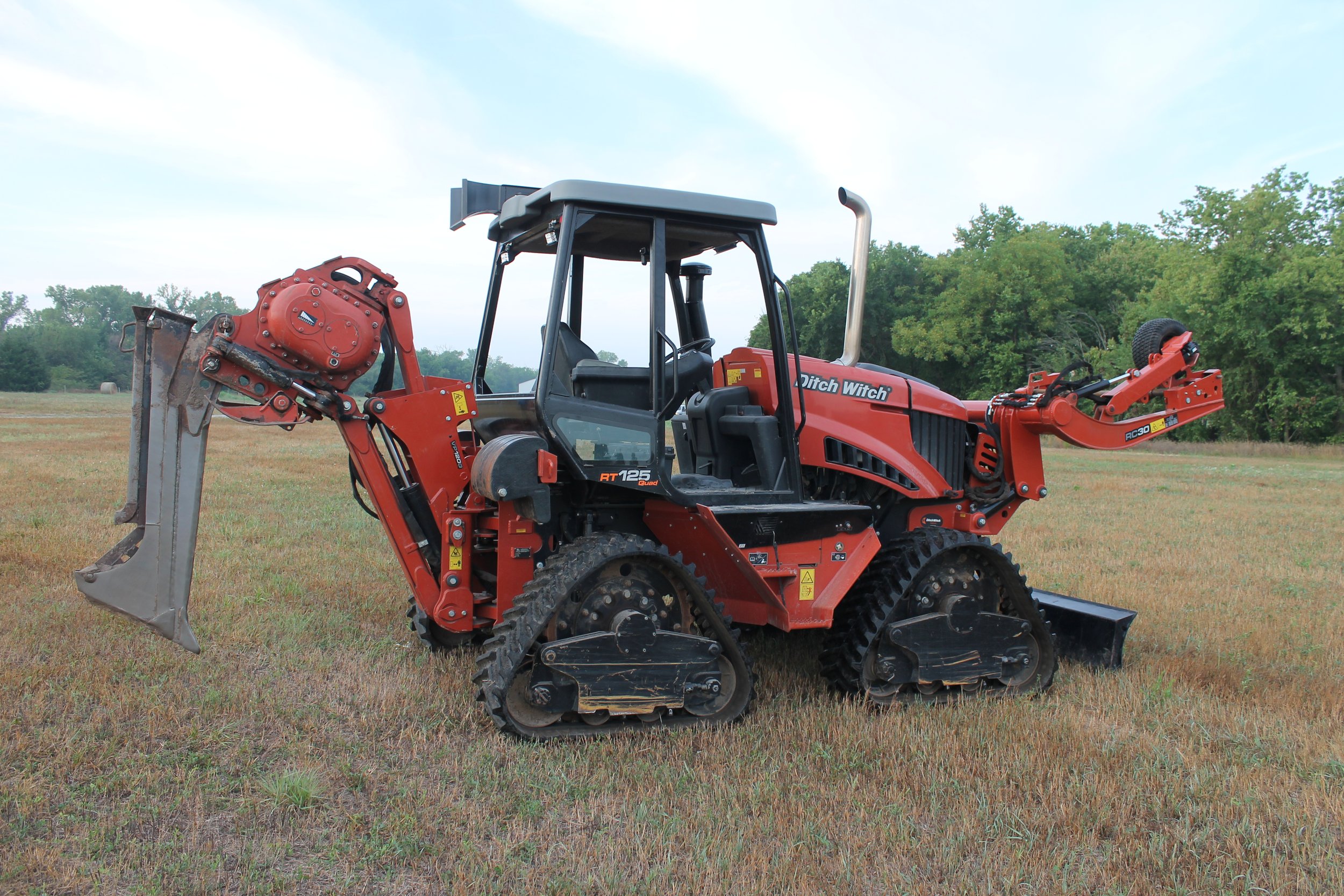 Red Ditch Witch RT125 Quad compact track loader with a front loader attachment on a grassy field, surrounded by trees under a partly cloudy sky.