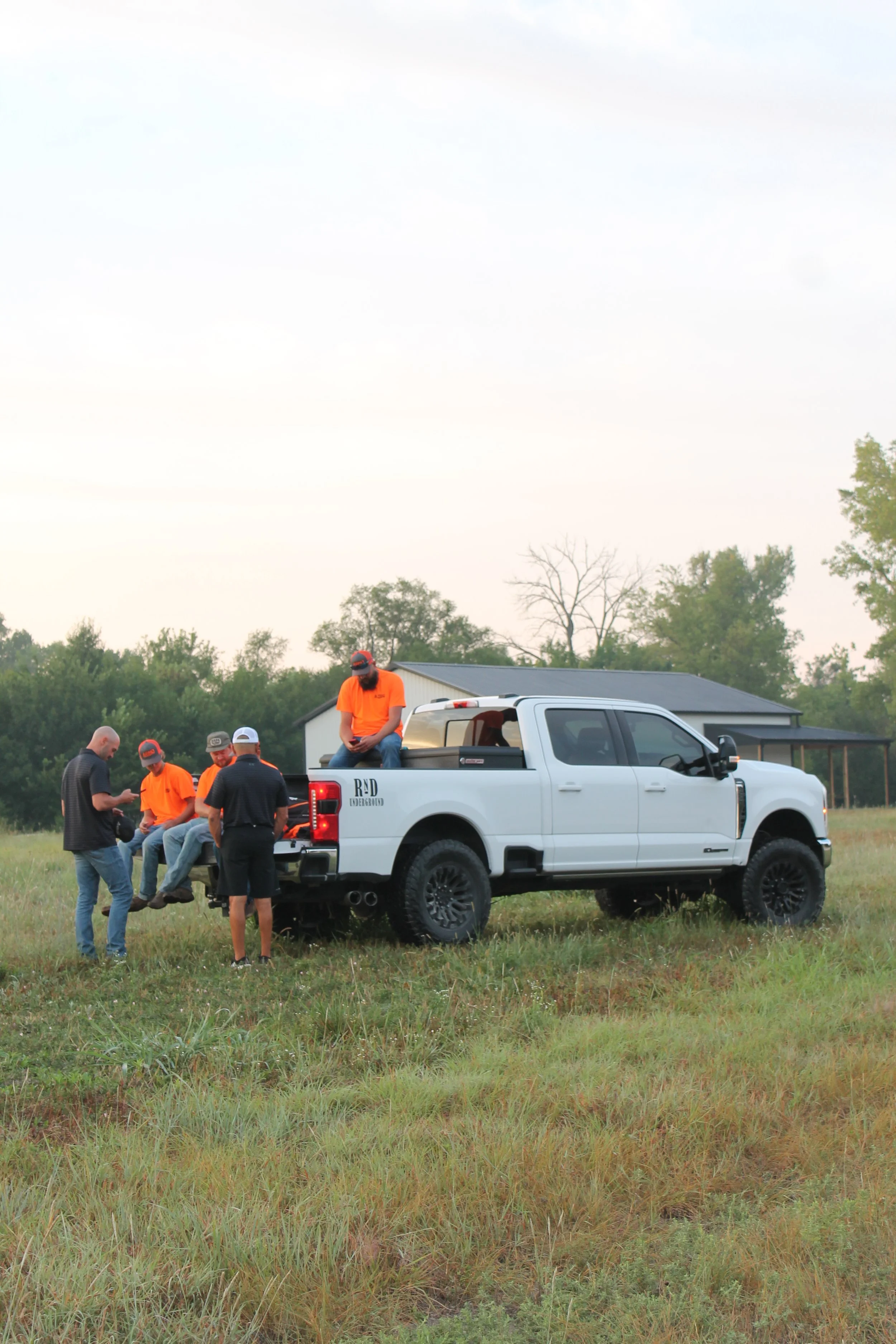 Group of people gathered around a white pickup truck in an open grassy field with trees and a building in the background, during daytime.