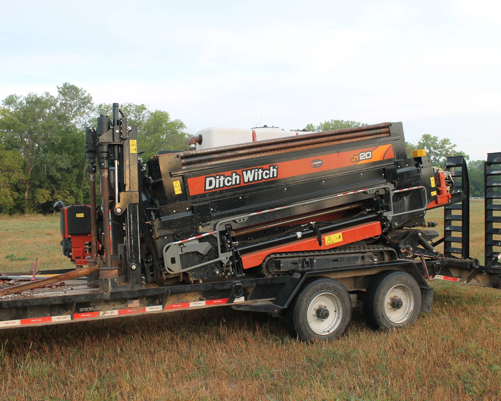 A flatbed truck carrying a Ditch Witch JT20 underground construction machine parked on grass with trees and a cloudy sky in the background.