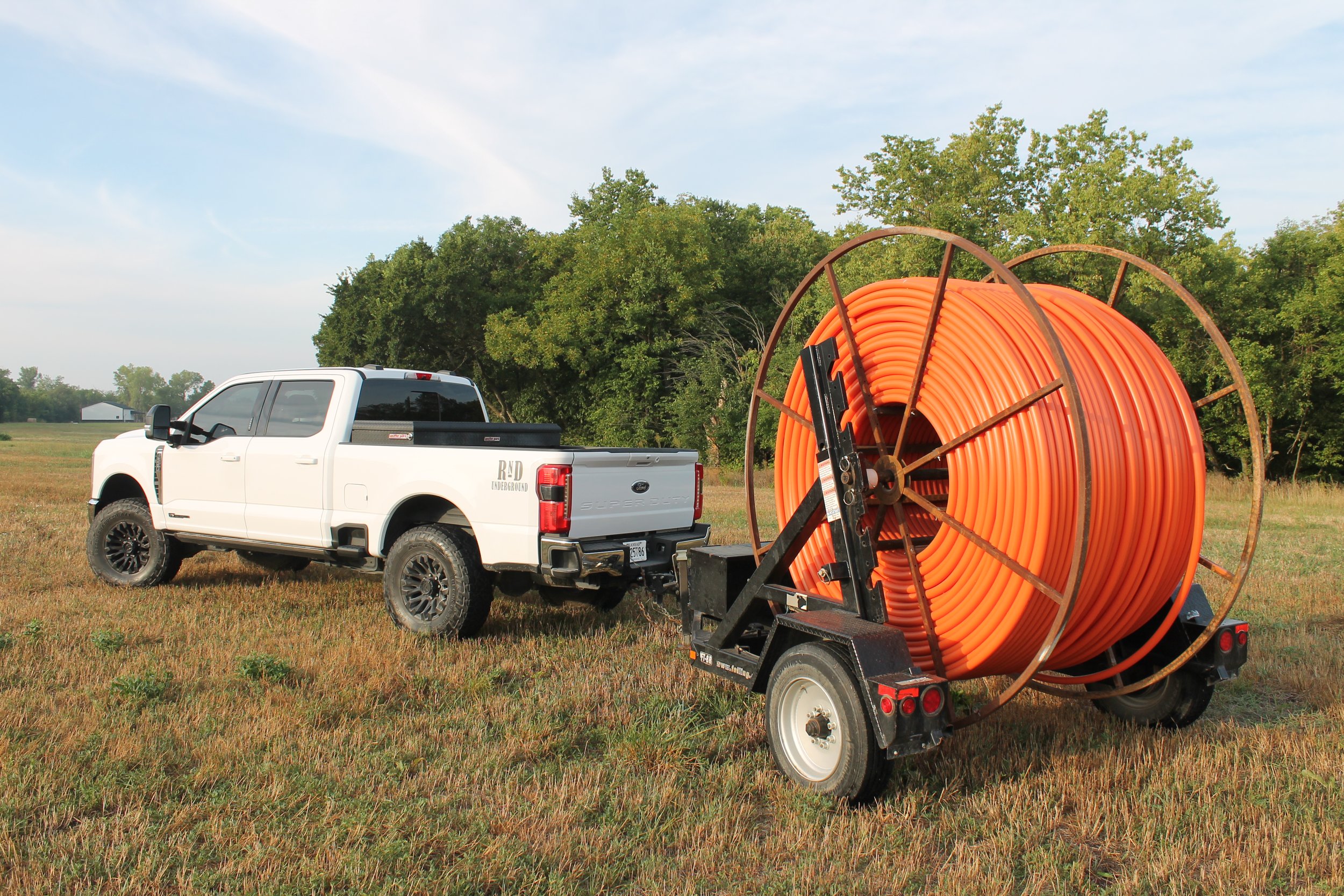 A white pickup truck parked on grass next to a large spool of orange piping mounted on a trailer, with green trees and blue sky in the background.