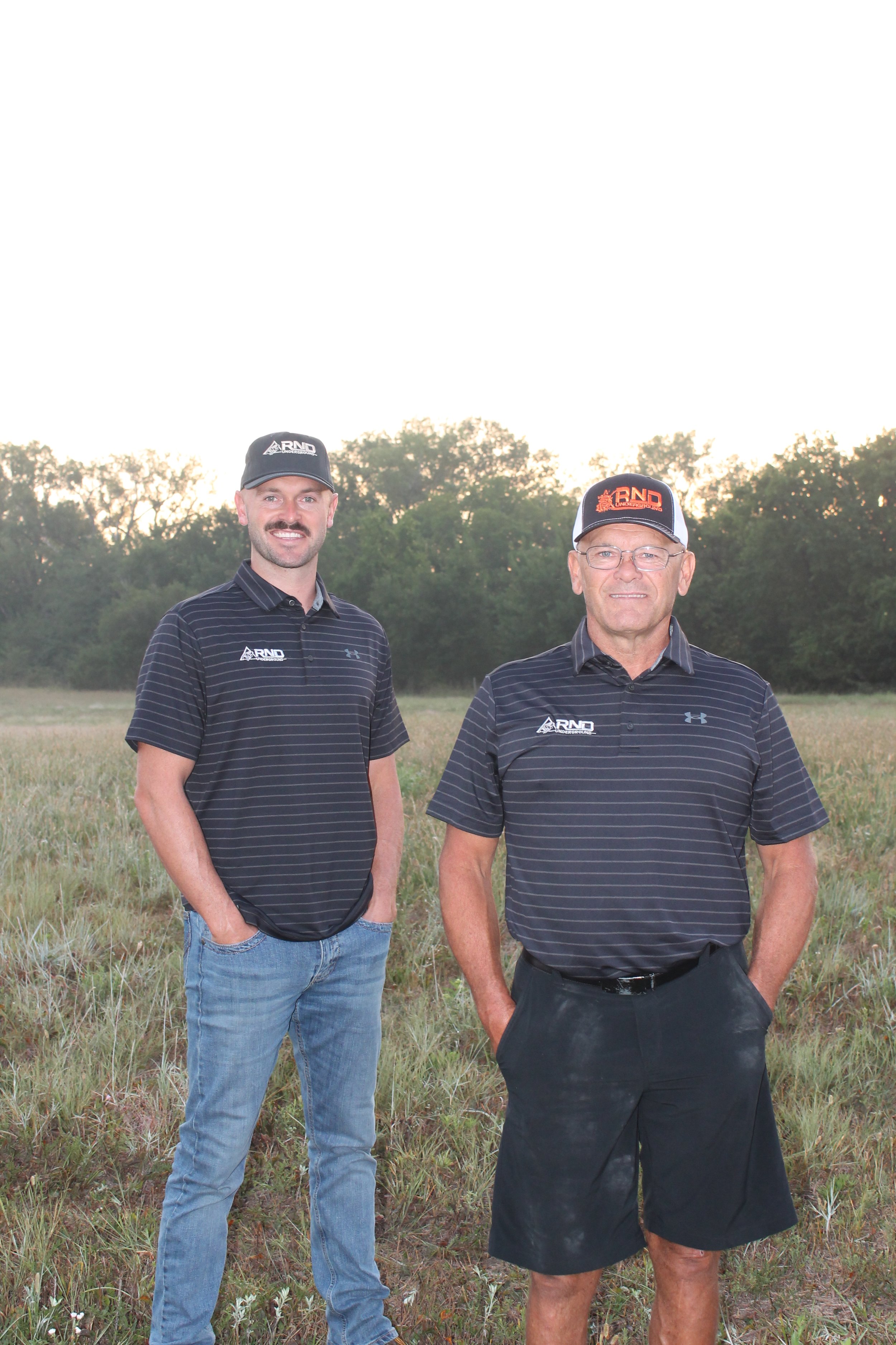 Two men standing outdoors in a grassy field with trees in the background, wearing black and white baseball caps and black striped polo shirts.