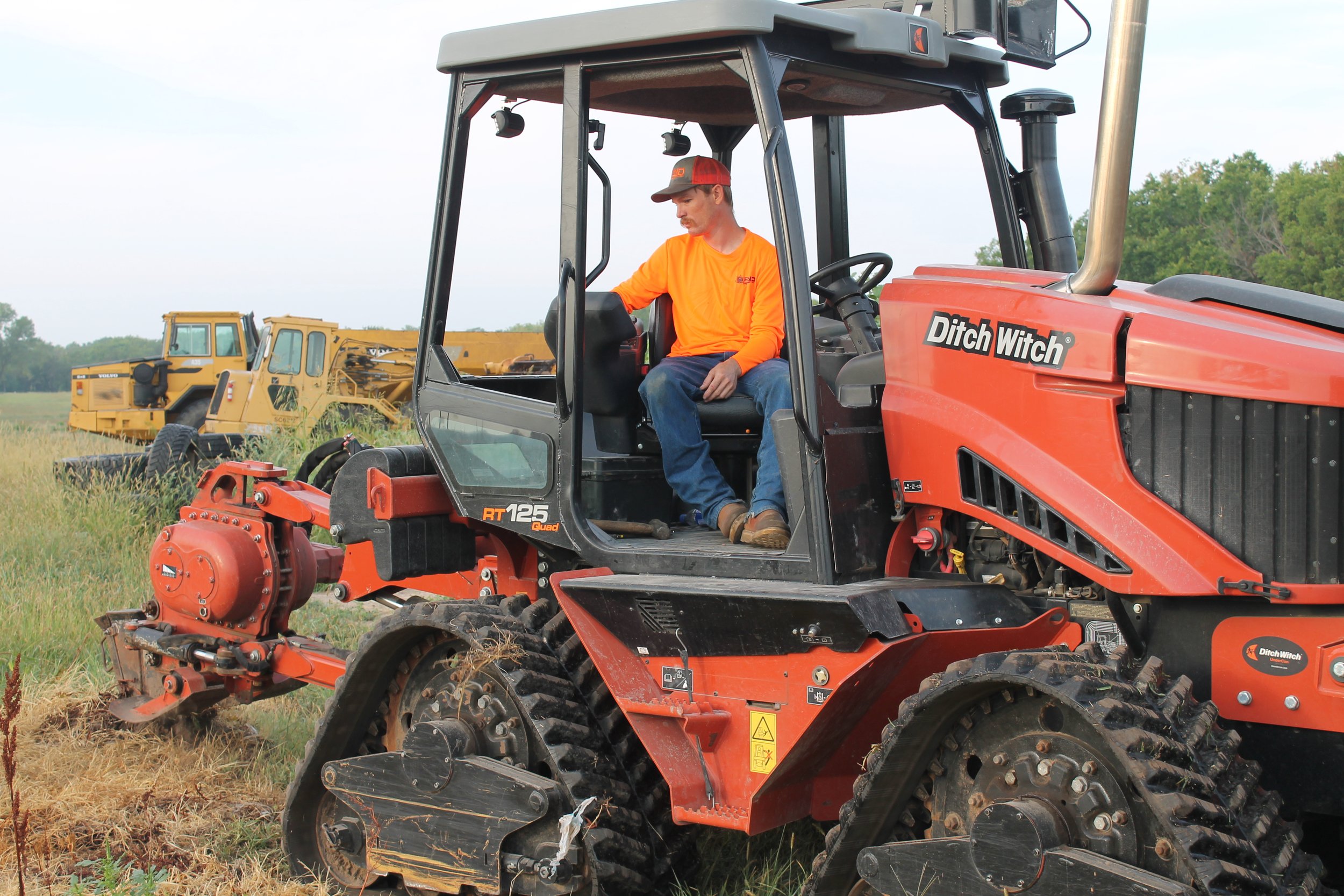 A man operating a Ditch Witch RT125 mini skid steer loader in a grassy field, with trees in the background.