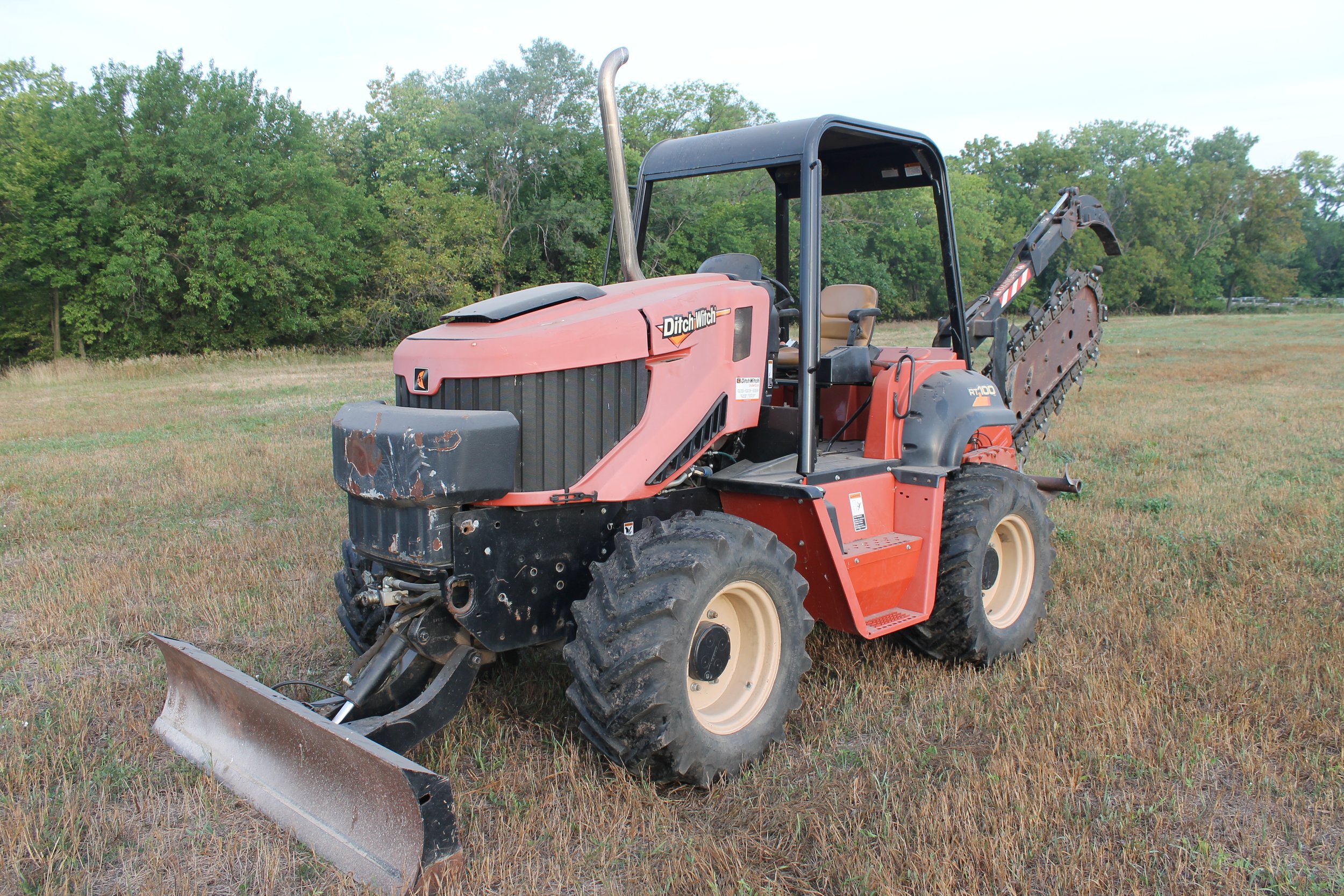 A red Ditch Witch trenching machine on a grassy field with a tree line in the background.
