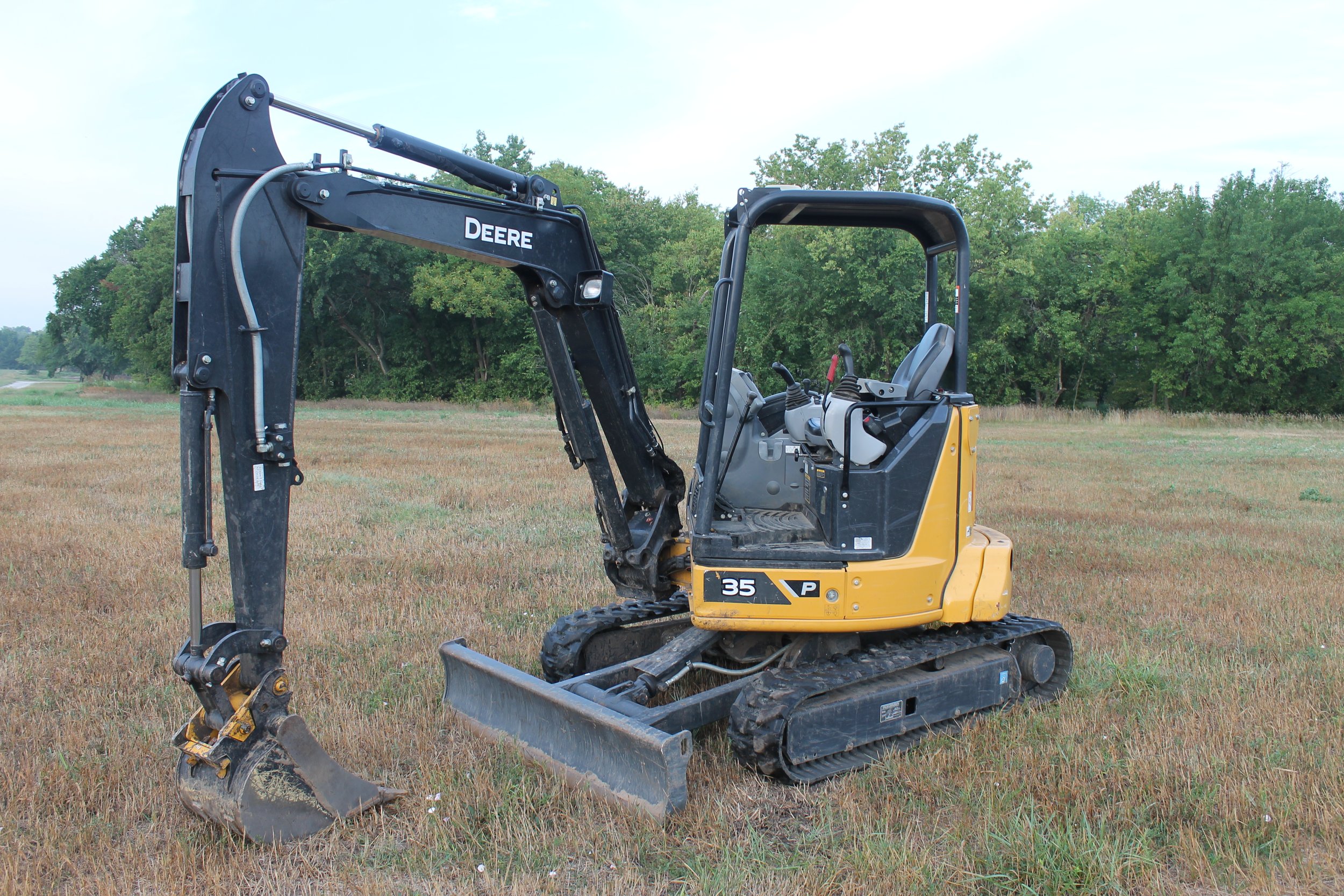 A yellow and black mini excavator with tracks, labeled "DEERE," parked on a grassy field with trees in the background.