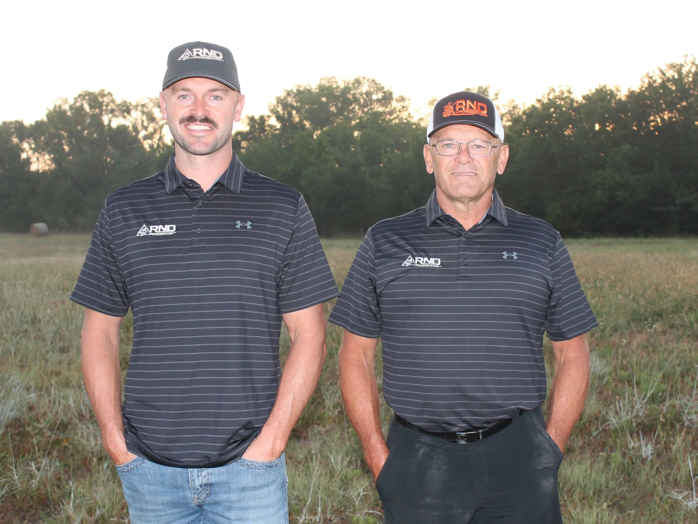 Two men standing outdoors in a grassy field with trees in the background, both wearing black and gray striped shirts and black caps with logos.