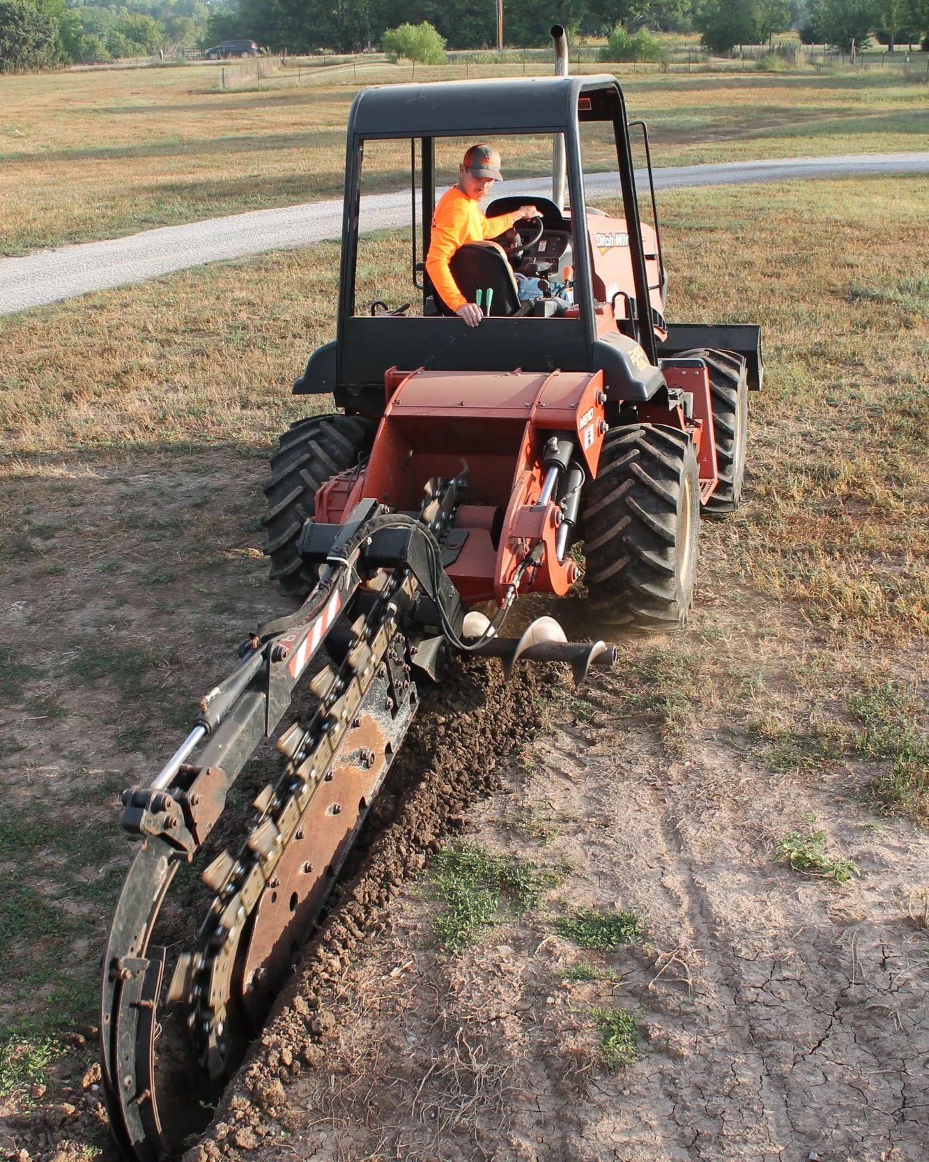 Young man driving a small black and orange tractor on a dirt field, wearing an orange shirt and a cap, surrounded by open land with a gravel road and trees in the background.