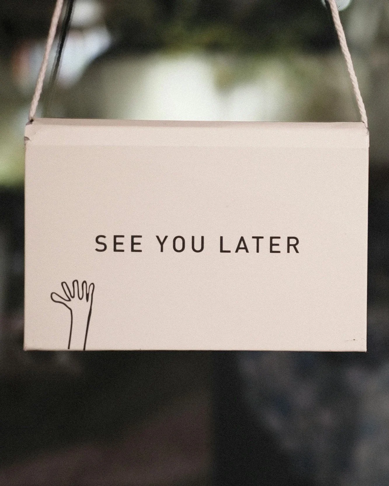 A white card with the words 'See You Later' printed on it, attached to a string, with a simple line drawing of a hand waving.