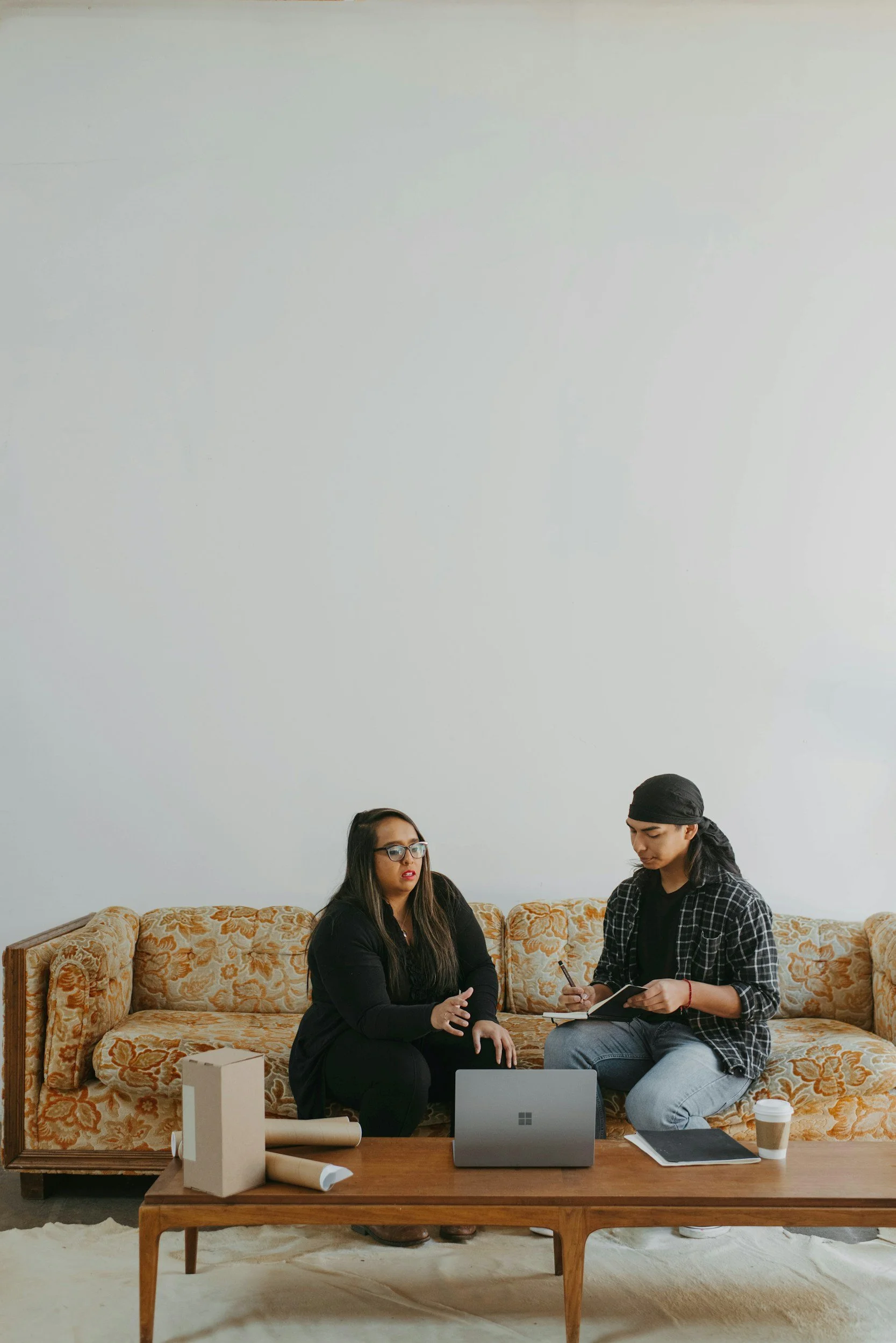 Two women are sitting on a floral-patterned vintage sofa in a casual office or creative space. One woman has long dark hair, glasses, and is wearing black. The other woman has dark hair under a black bandana, and is wearing a black plaid shirt, holding a notebook and pen. In front of them, there is a wooden table with a Microsoft Surface laptop, some rolled papers, a white box, and a disposable coffee cup. Behind them, a plain white wall and a beige carpet are visible.