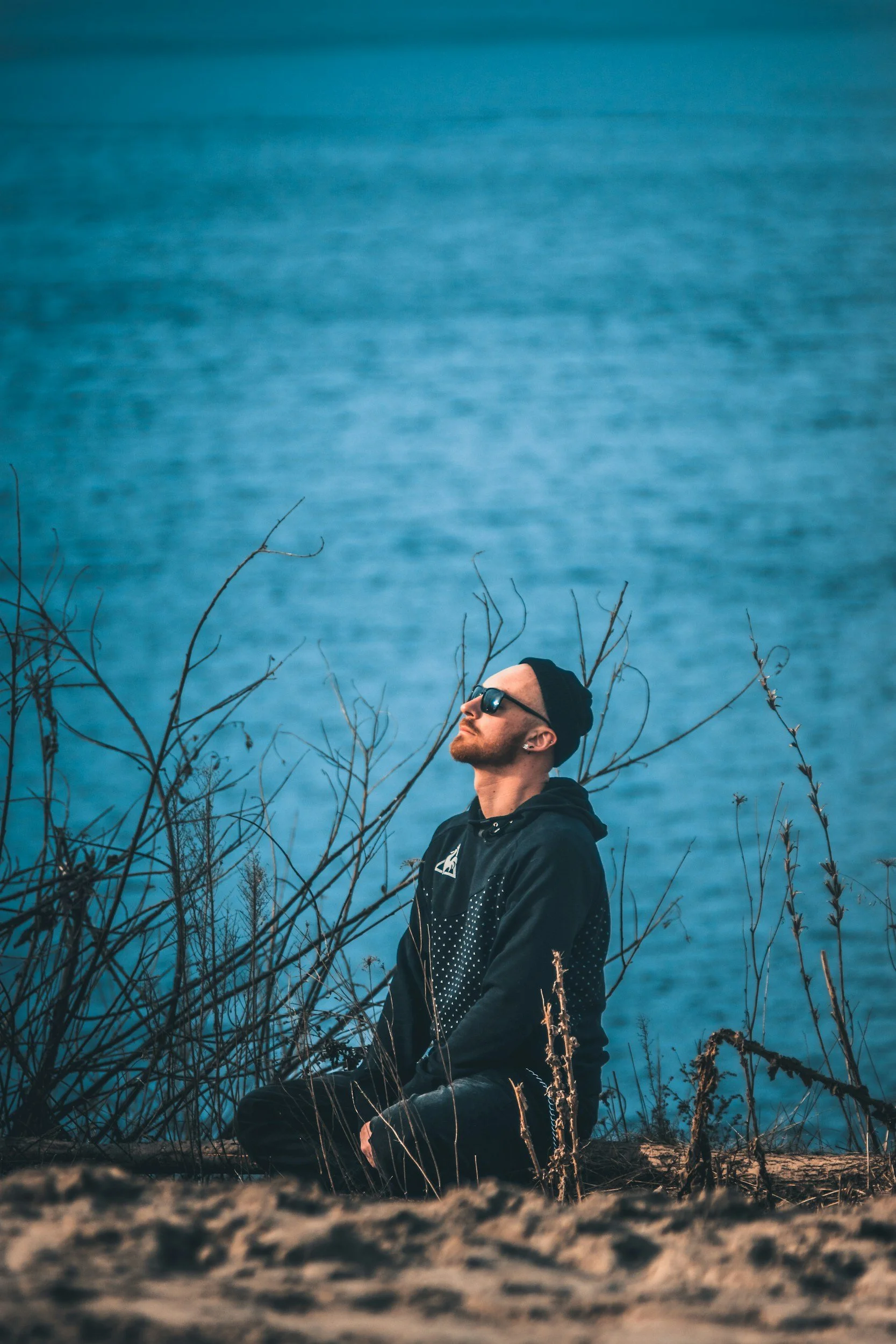 A man wearing sunglasses and a black hoodie with white dots sitting outdoors amidst leafless branches, with a large body of water in the background, under an overcast sky.