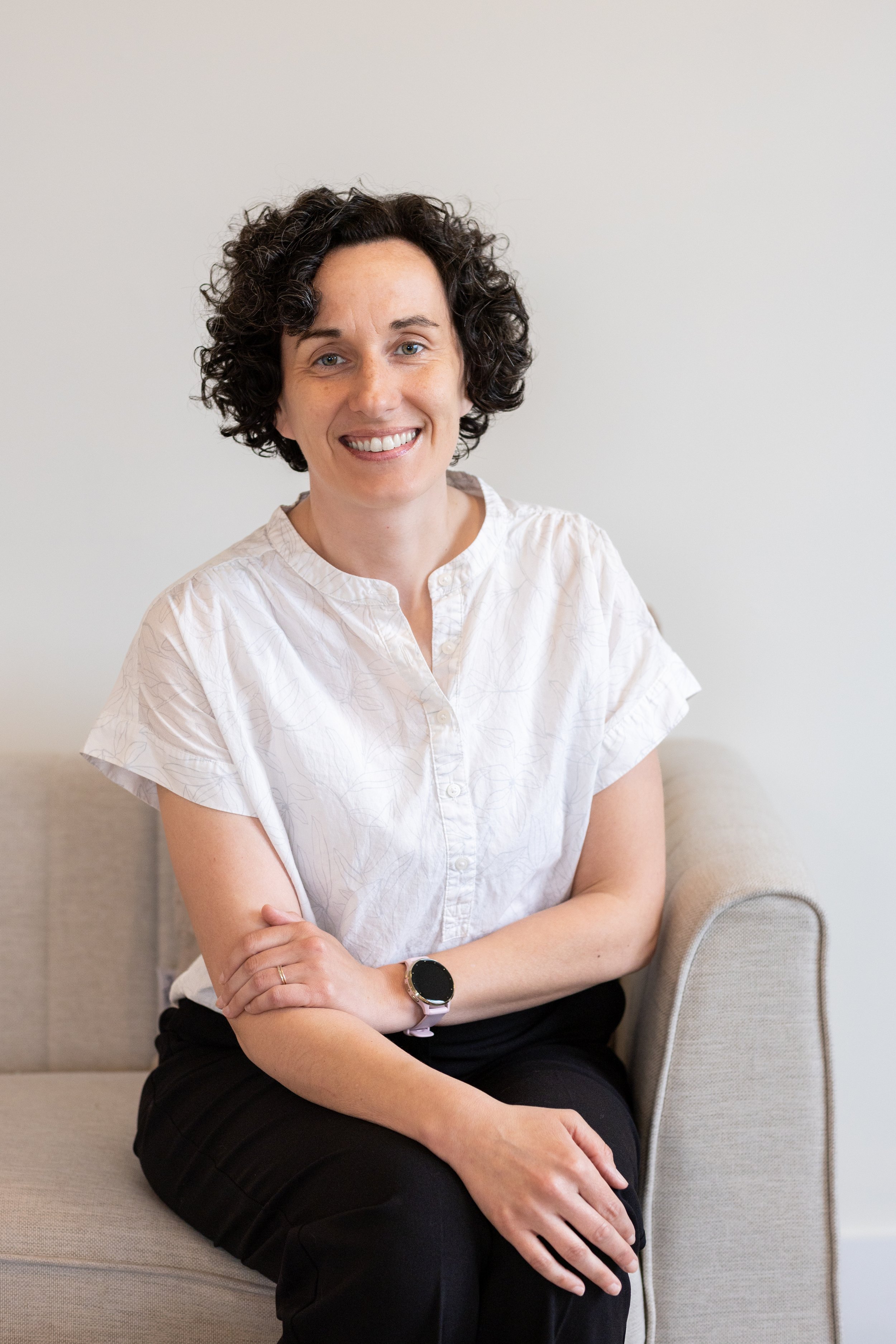 A woman with curly dark hair, wearing a white blouse and black pants, sitting on a beige sofa with her arms crossed, smiling at the camera.