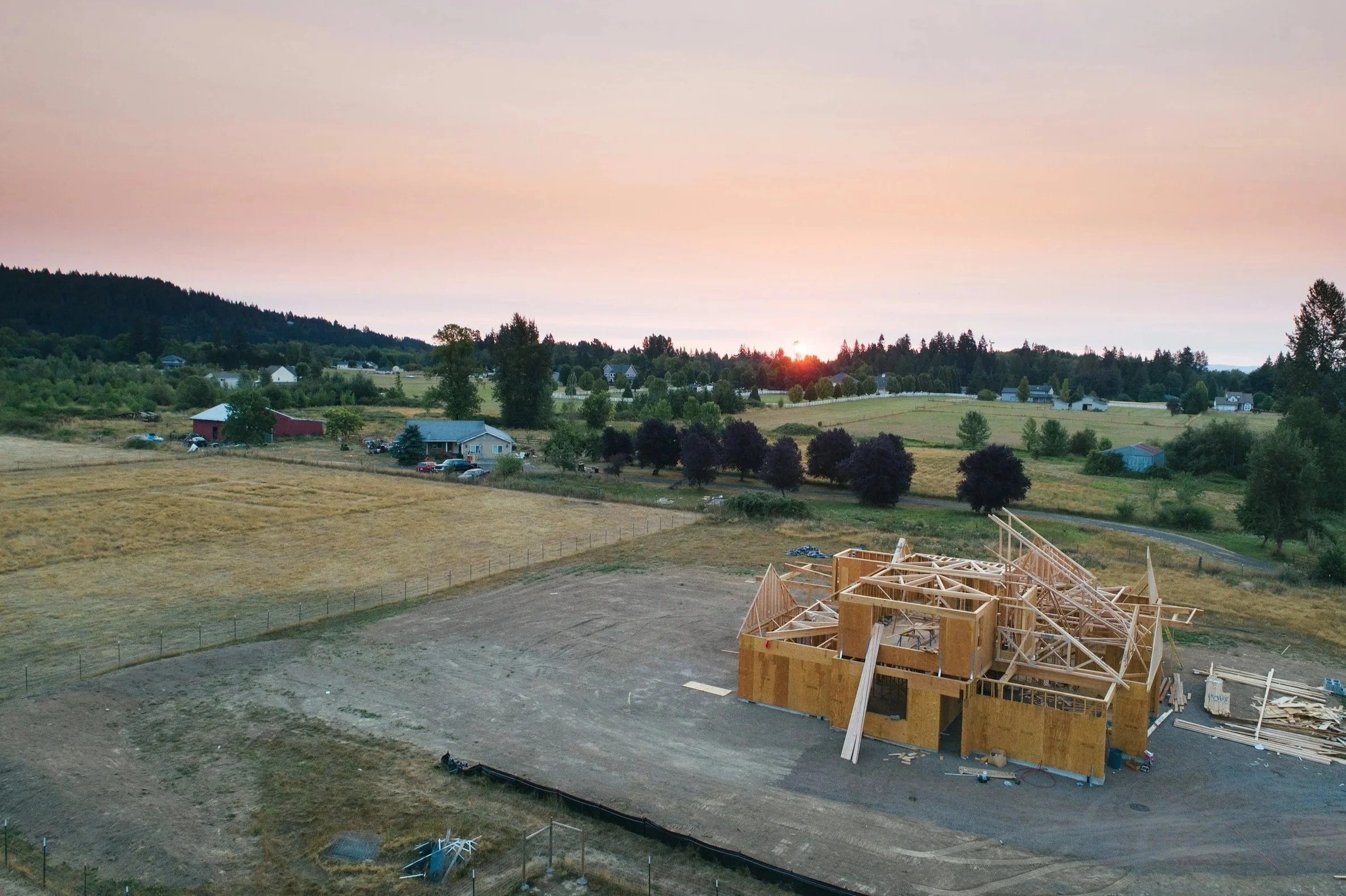 A construction site with unfinished wooden framing for a building, set against a rural landscape during sunset with houses, trees, and rolling hills in the background.
