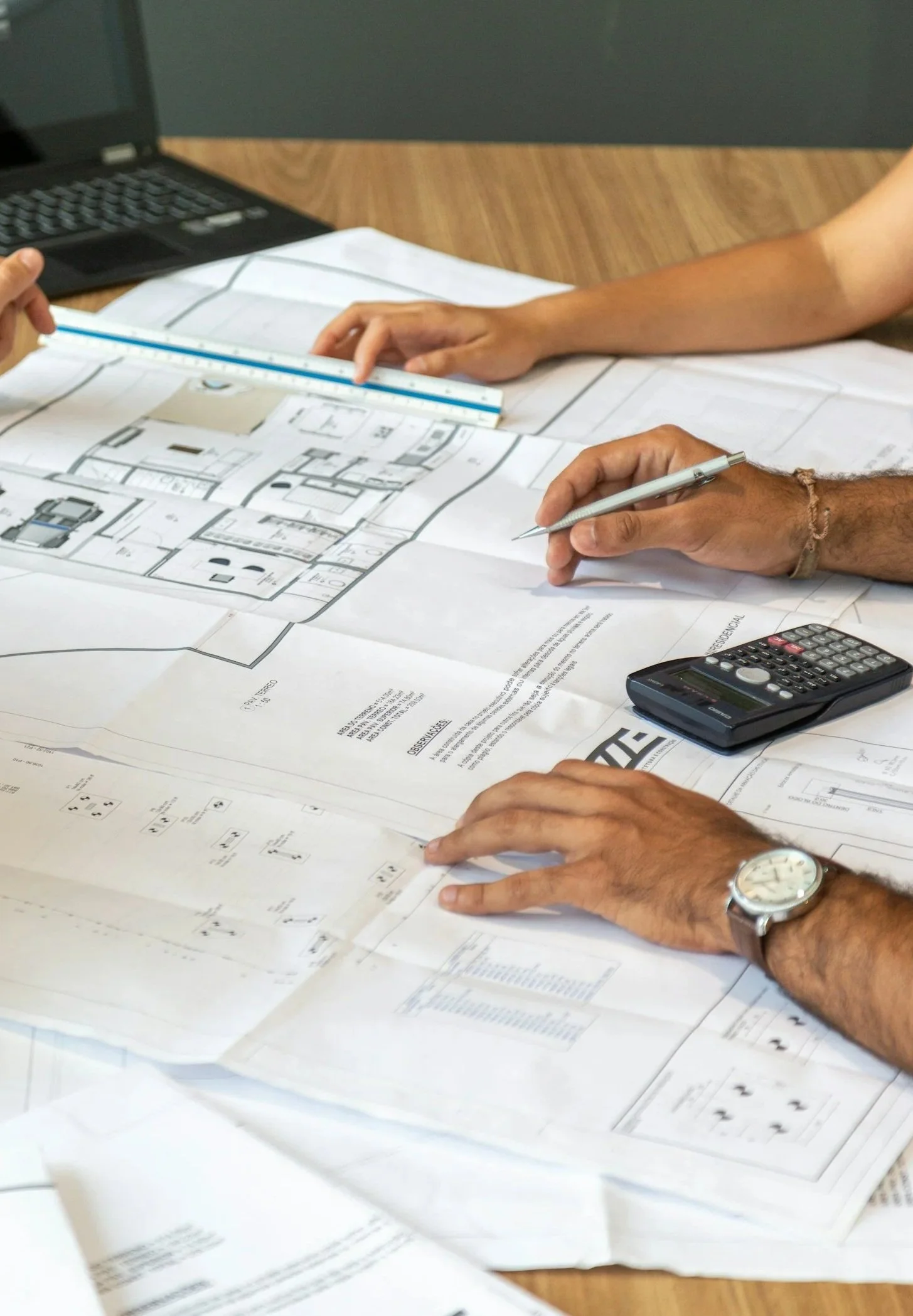 Two people reviewing architectural floor plans on a wooden table, with a laptop, calculator, and writing tools.