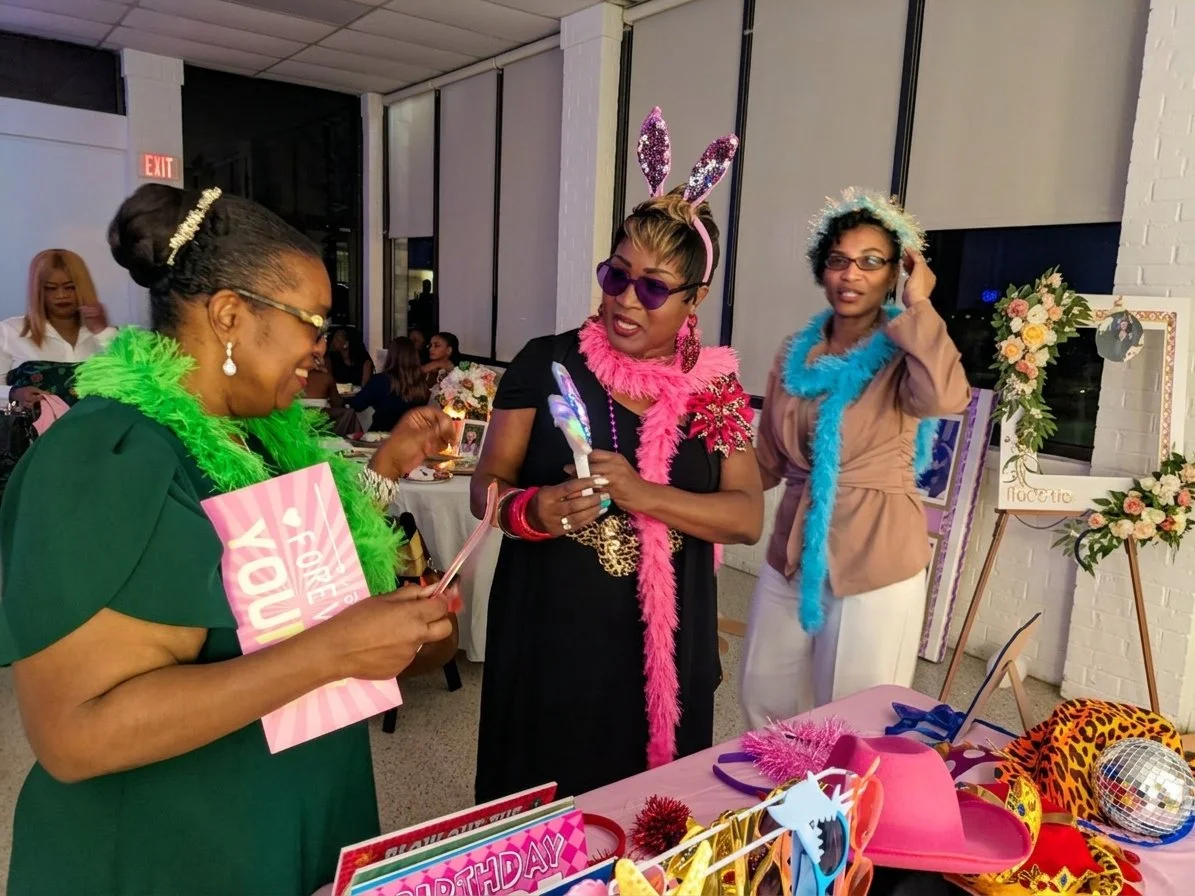Three women wearing colorful feather boas and headbands, engaging at a birthday celebration with pink party items, flowers, and decorations.