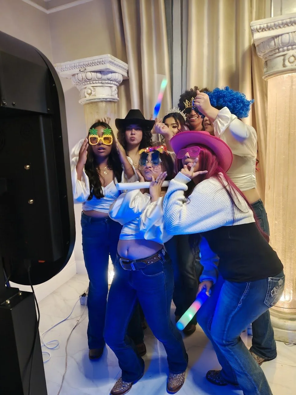 Group of seven women dressed in costume, wearing silly glasses, hats, and masks, posing playfully in front of decorative columns and curtains at a party.