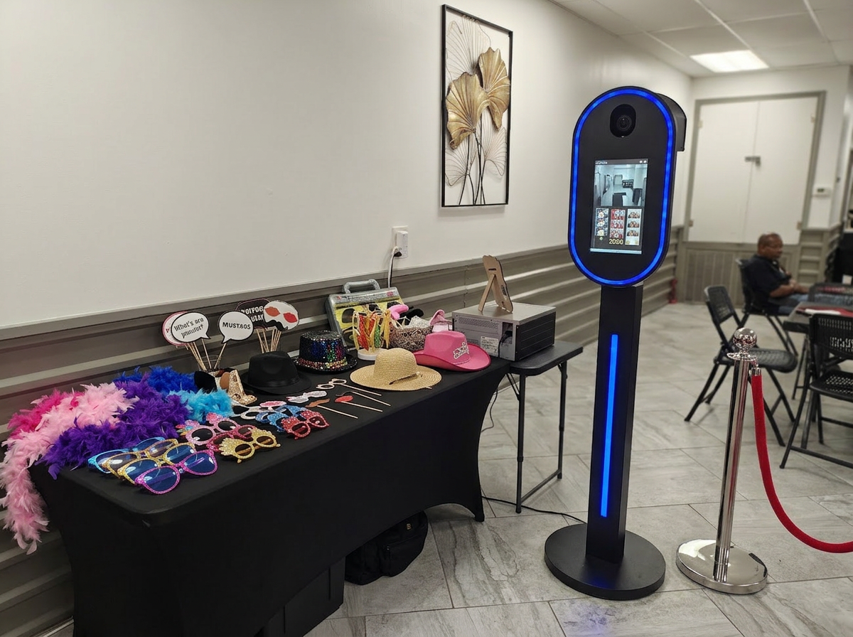 Photobooth setup with various props including feather boas, sunglasses, hats, and signs, next to a digital photobooth kiosk in an indoor setting.