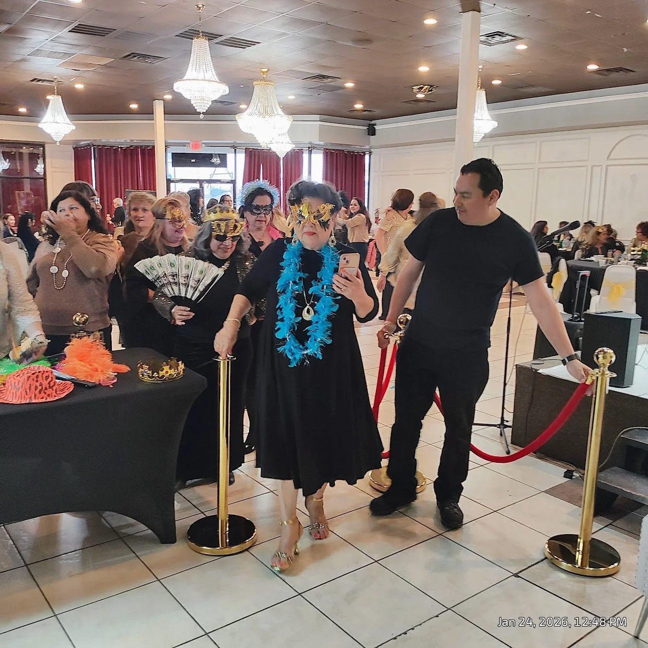 Group of people dressed in costumes at a festive indoor event, with some wearing masks, feather boas, and holding props, gathered around a table with colorful items and accessories, in a decorated venue with chandeliers and red curtains.