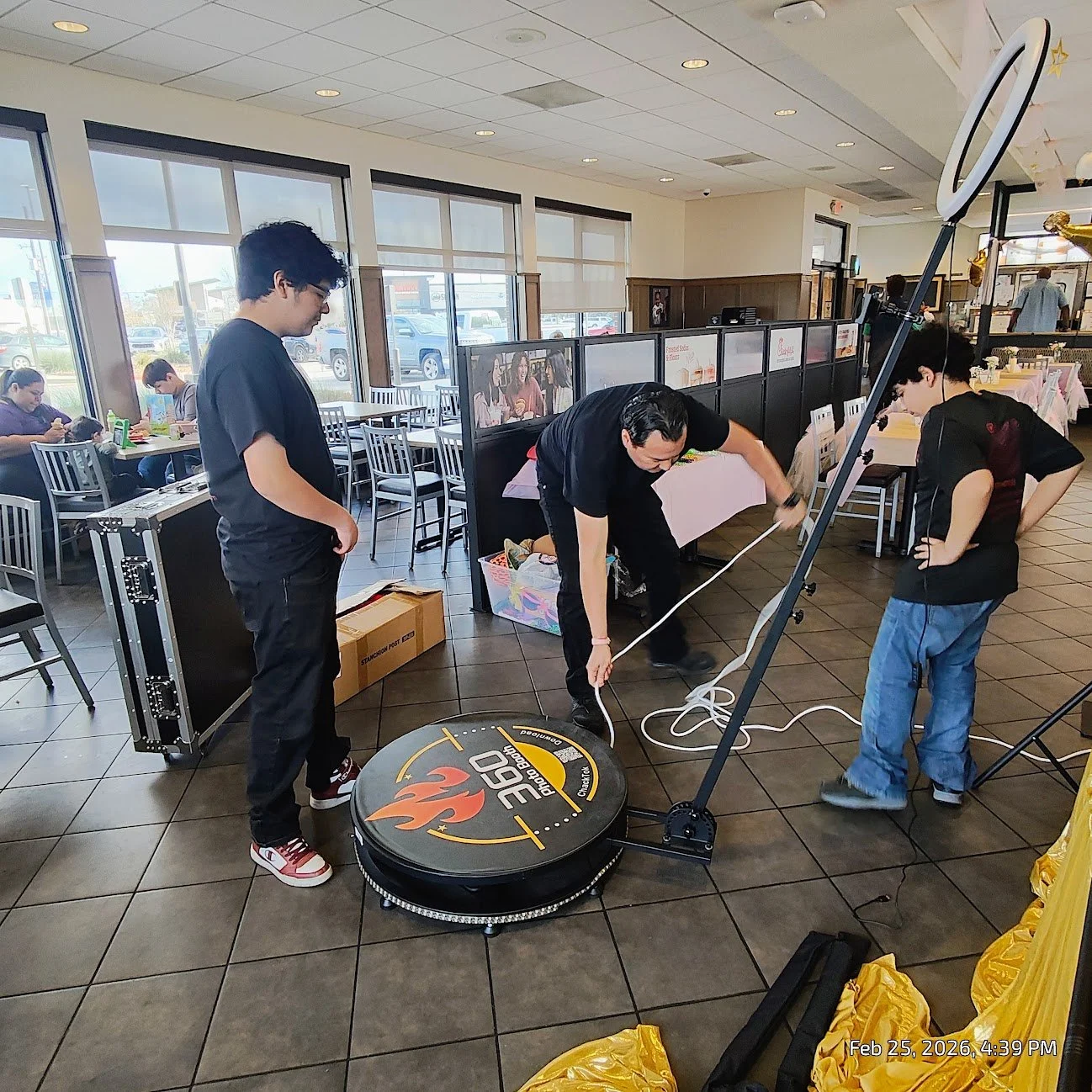 Three men are setting up equipment inside a restaurant. One man is working with wires near a robotic device on the floor. The other two men are observing. The restaurant has tables, chairs, and large windows with natural light. Some people are seated