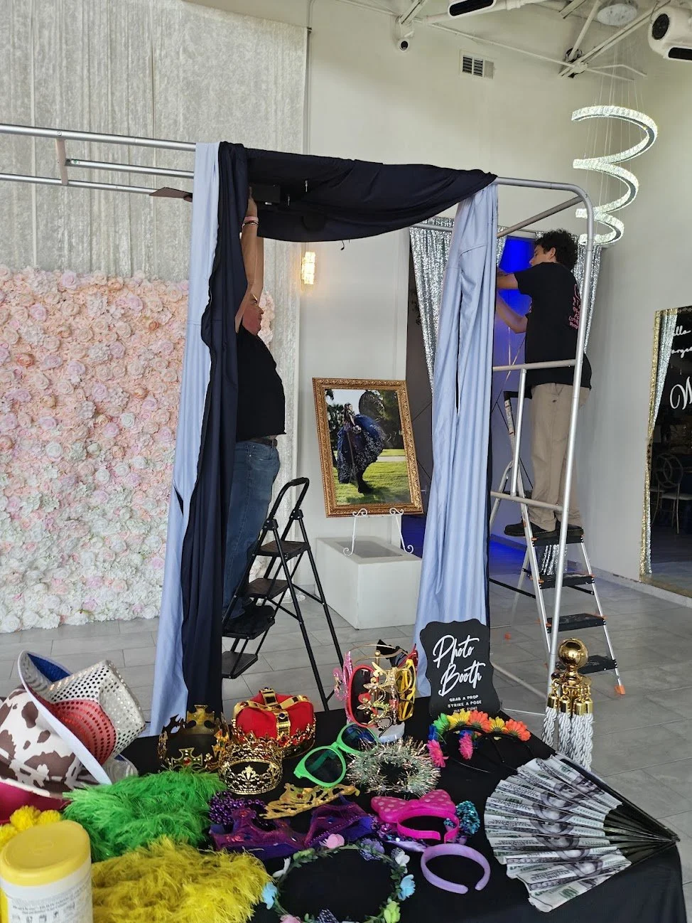 Setup for a photo booth with decorations, props like crowns, hats, glasses, and a floral backdrop, with workers preparing the area inside a decorated event space.