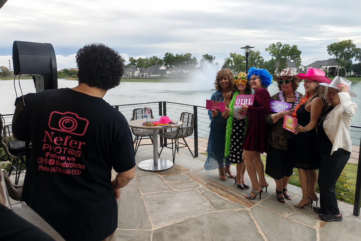 Group of women dressed in fun costumes and accessories standing on a patio near a lake, posing for a photo while a photographer takes their picture. The women are holding colorful signs and wearing wigs, hats, and sunglasses.
