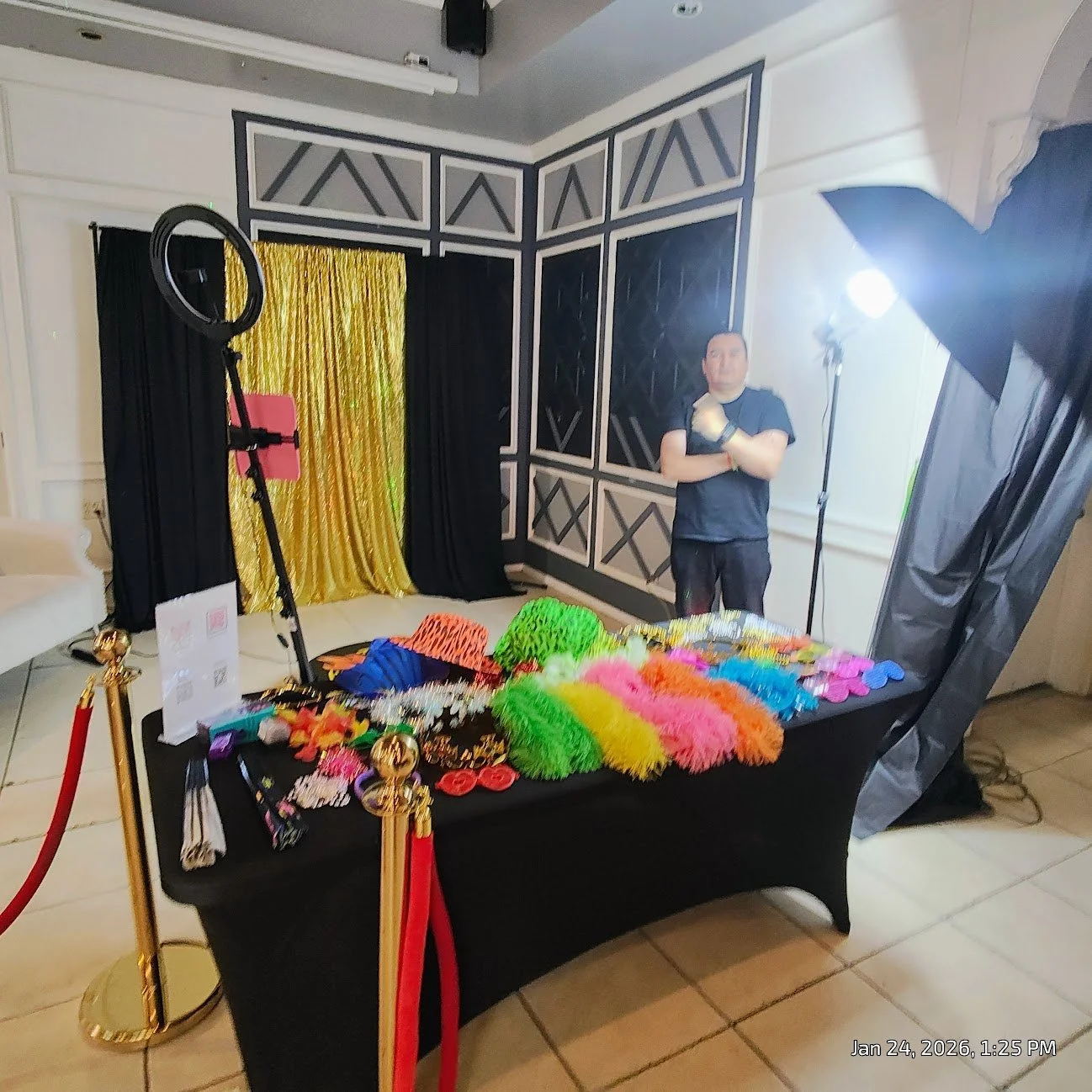 Photo of a photo booth setup with colorful decorations and accessories, a man standing behind the table, and professional lighting equipment in a room with black, white, and gray walls.