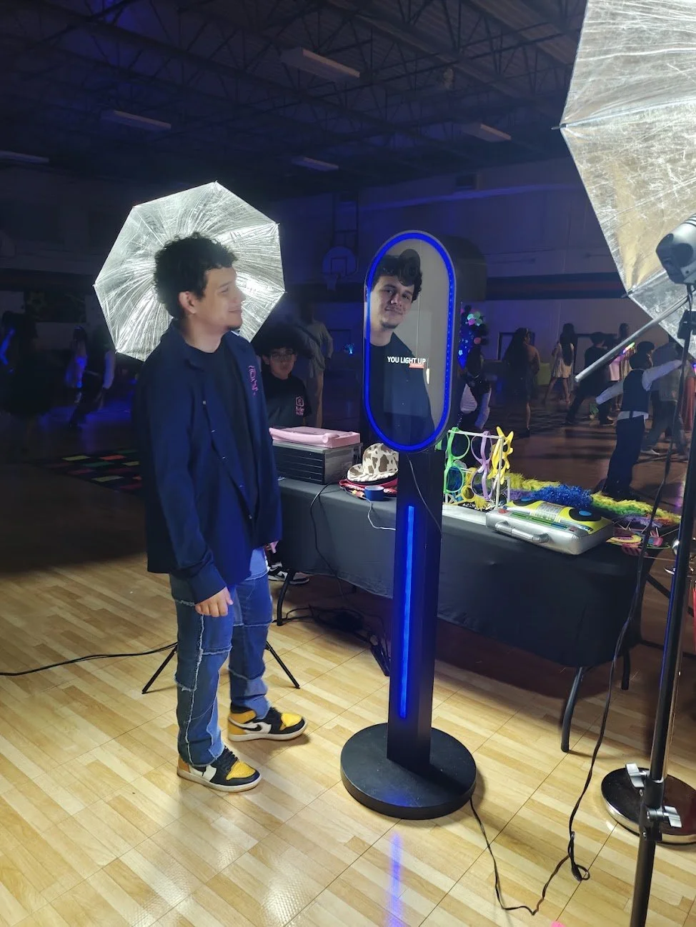 A young man with curly black hair and a navy blue jacket stands in front of a light-up digital mirror at a dance or party event. The area is dimly lit with other people dancing in the background. There are various electronic equipment and colorful ac
