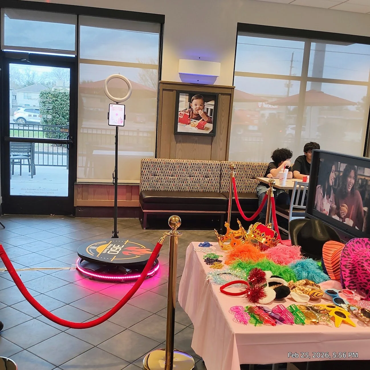 Inside a restaurant decorated for a birthday party with a red velvet rope barrier, a table with colorful costumes and accessories, a photo booth with a ring light, and a family enjoying their meal.