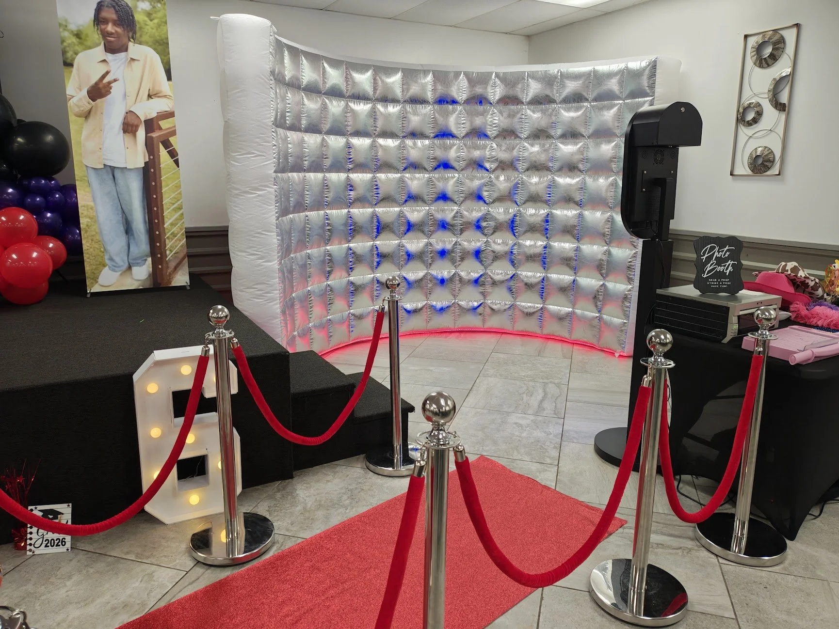 Photo booth area with a red carpet and velvet ropes, a metallic backdrop, a sign that says 'Photo Booth,' and props on a table to the right. To the left is a cardboard cutout of a person making a peace sign.