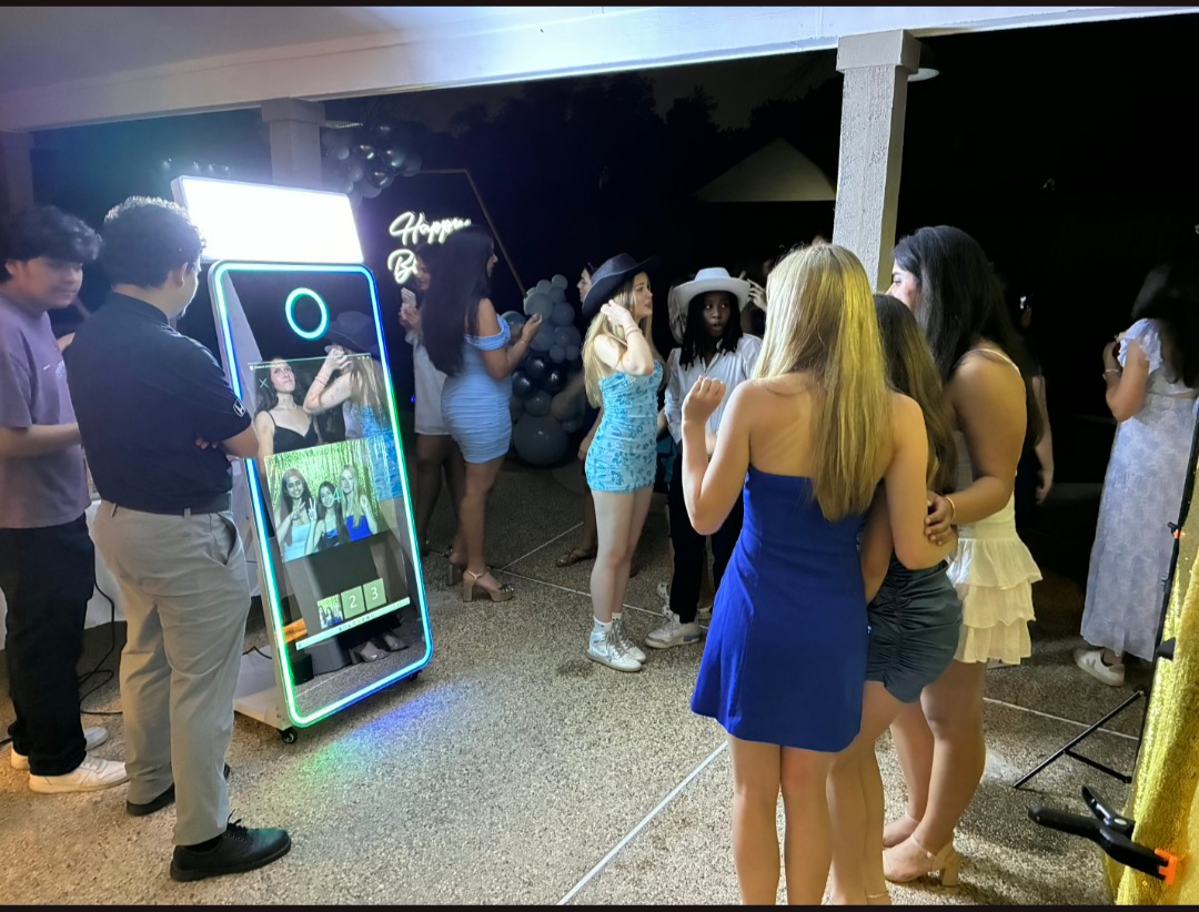 People gathered around a photo booth at a party, with some posing and others waiting, decorated with balloons and a neon sign that reads 'Happy B'.