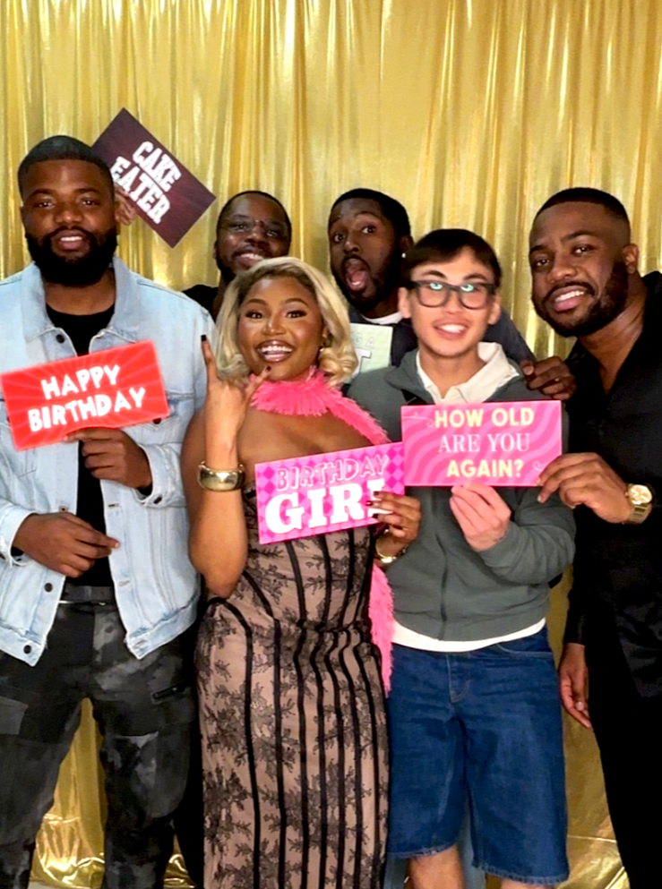 Group of friends celebrating a birthday, holding colorful signs that say "Happy Birthday," "Birthday Girl," and "How Old Are You Again?" in front of gold curtain backdrop.