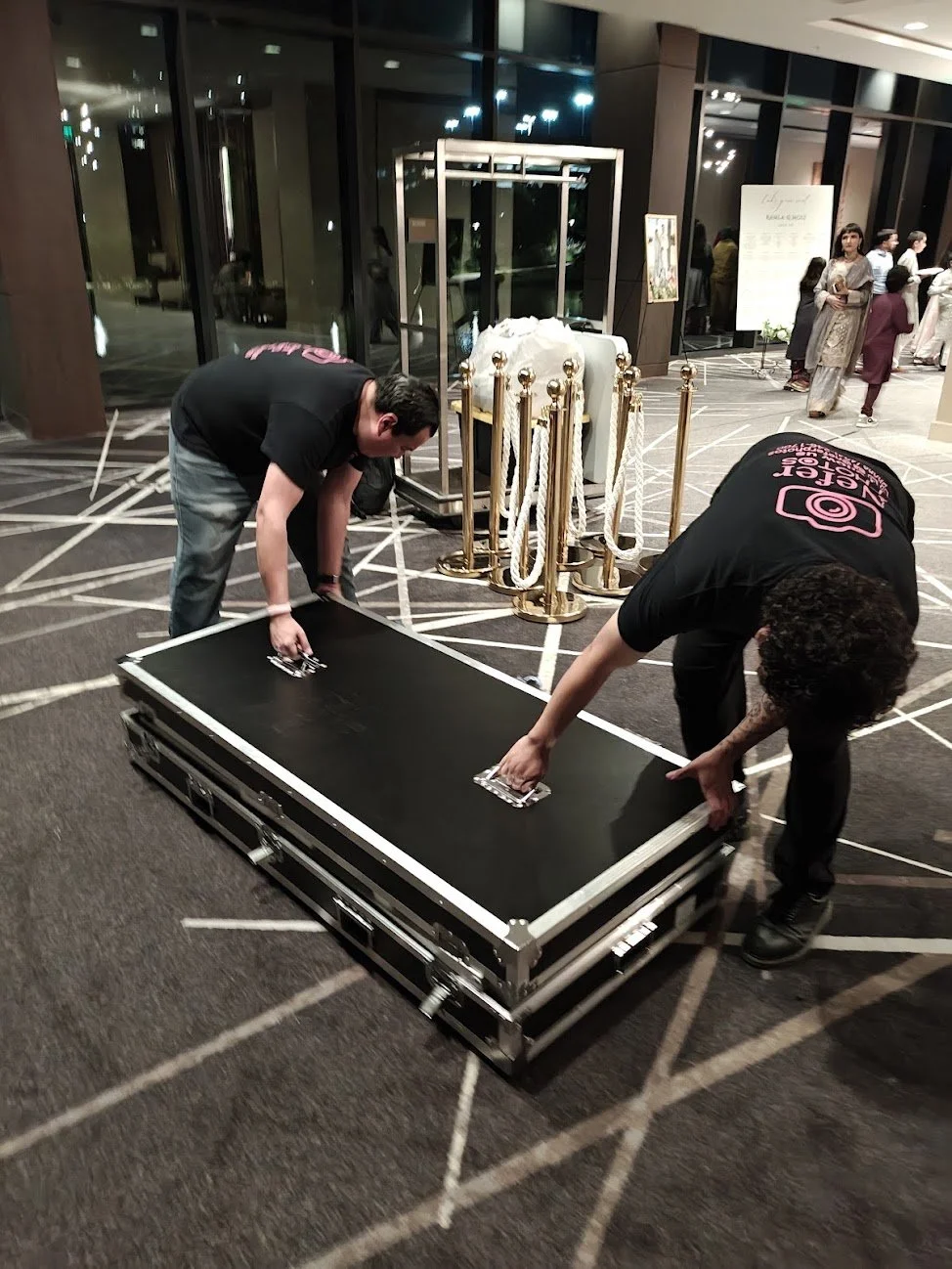 Two people are setting up or packing a large black case on the floor in a hotel lobby, with event attendees in the background.
