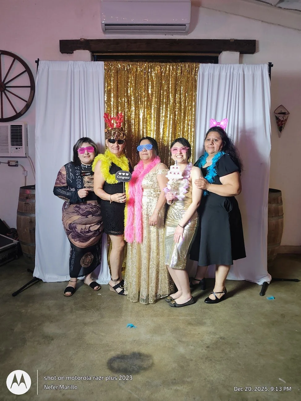 Group of five women celebrating with birthday props in front of a gold sequin backdrop and white curtains, wearing colorful glasses and feather boas.