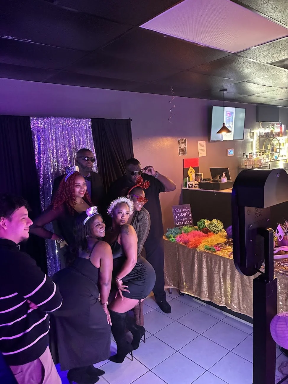 A group of people dressed up in party attire, posing together at a celebration or party in a decorated room with a sequin backdrop, a table with colorful accessories, and a bar area in the background.