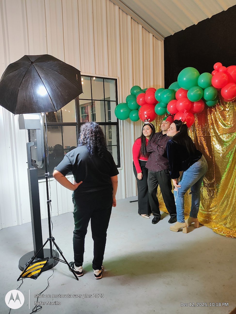 People posing for a photo at a holiday-themed photo booth with a golden curtain backdrop and green and red balloons. A photographer is taking their picture using a studio light with an umbrella. The photo is taken indoors, near a window with a beige 