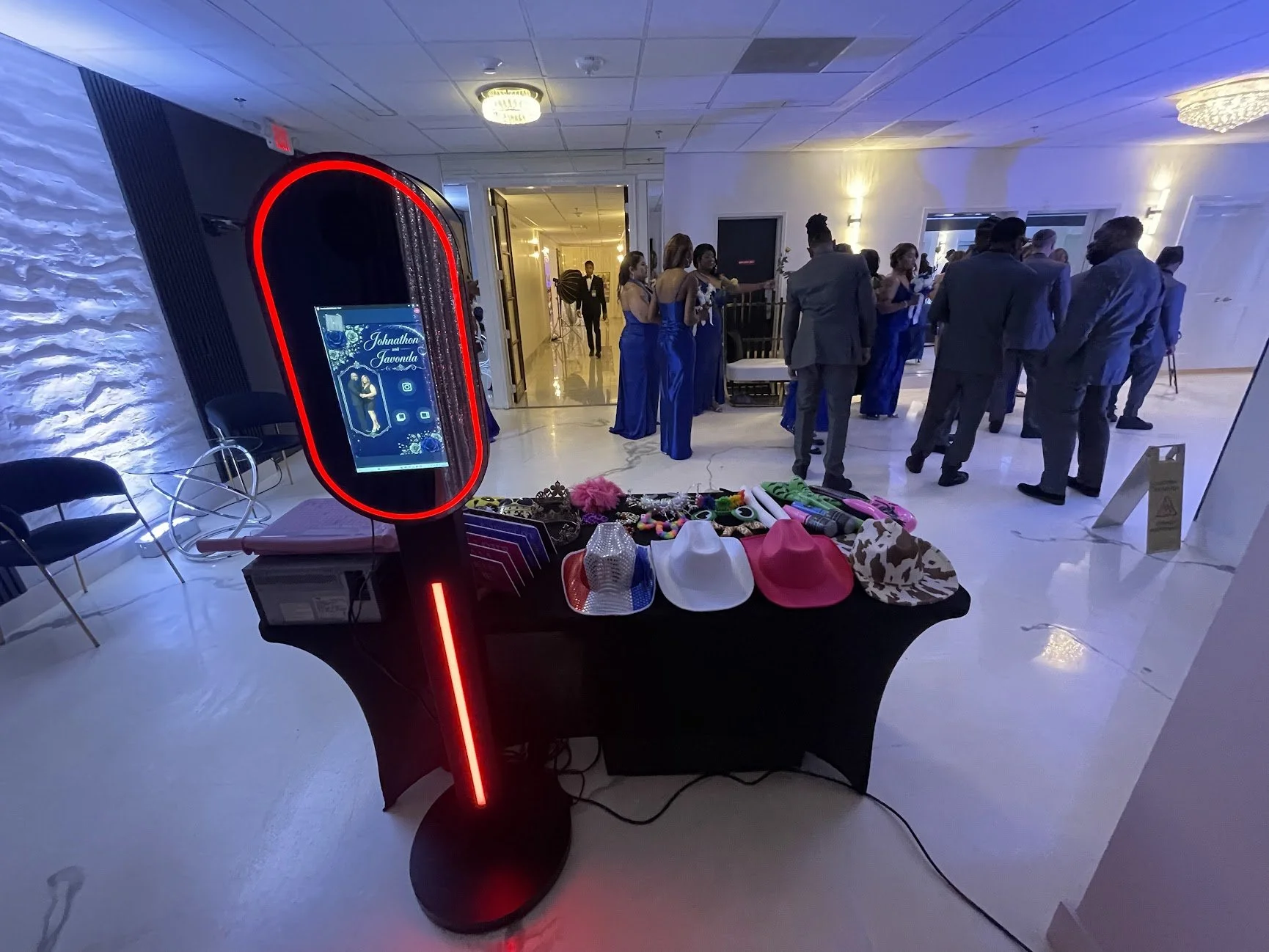 A photo of a social gathering in a banquet hall. In the foreground, a table displays various hats and accessories. To the left, a digital photo booth with the names 'Johnathon & Javerda' on the screen. In the background, people dressed in formal atti