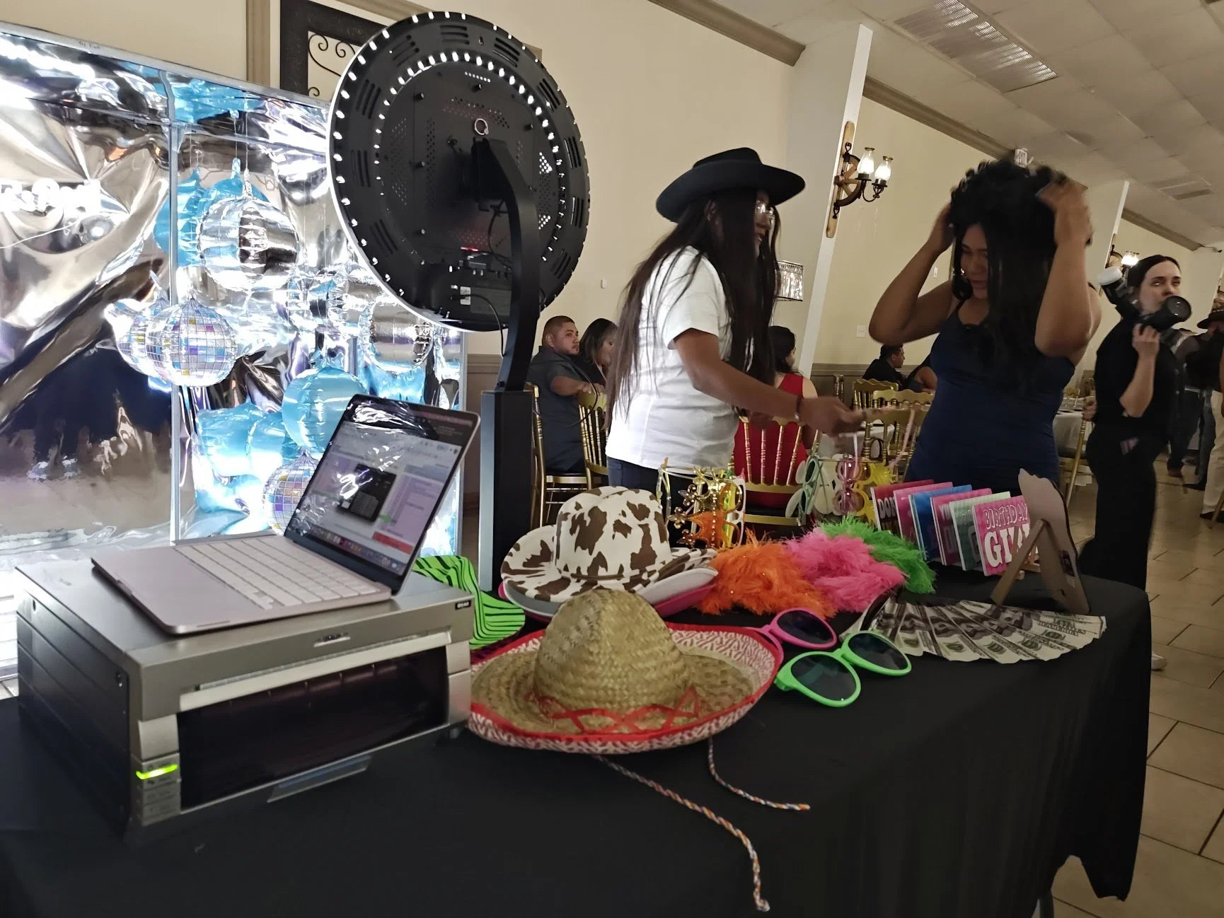 A decorated table at a party or event with various accessories including hats, sunglasses, and novelty items. There is a laptop on a corner of the table, and people are dancing and socializing in the background.