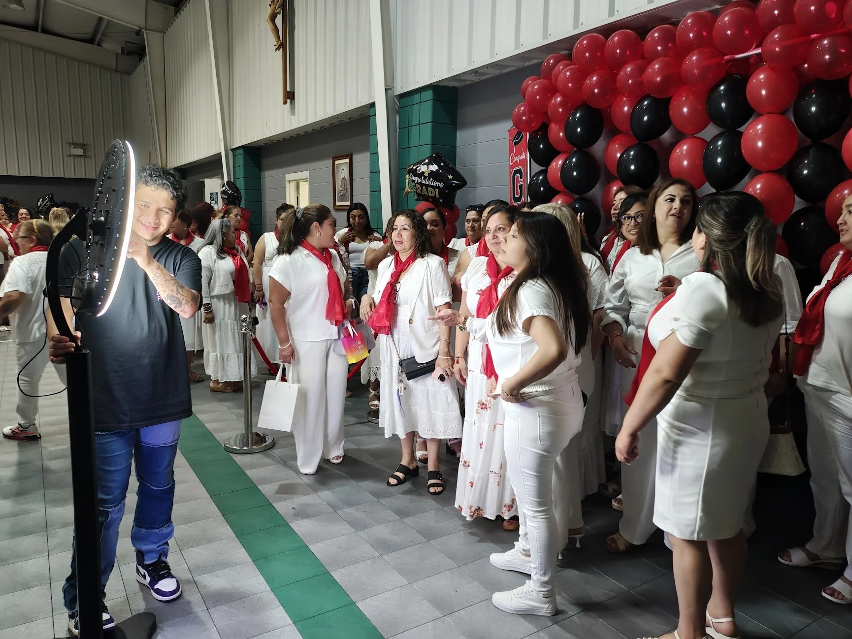 Group of women dressed in white, gathered at a celebration with red and black balloons and a portrait of Jesus Christ on the wall.