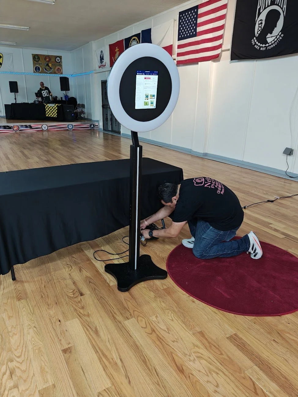 A man kneeling on the floor adjusting a selfie station with a built-in screen in an event hall decorated with American flags, military flags, and a stage with DJ equipment in the background.