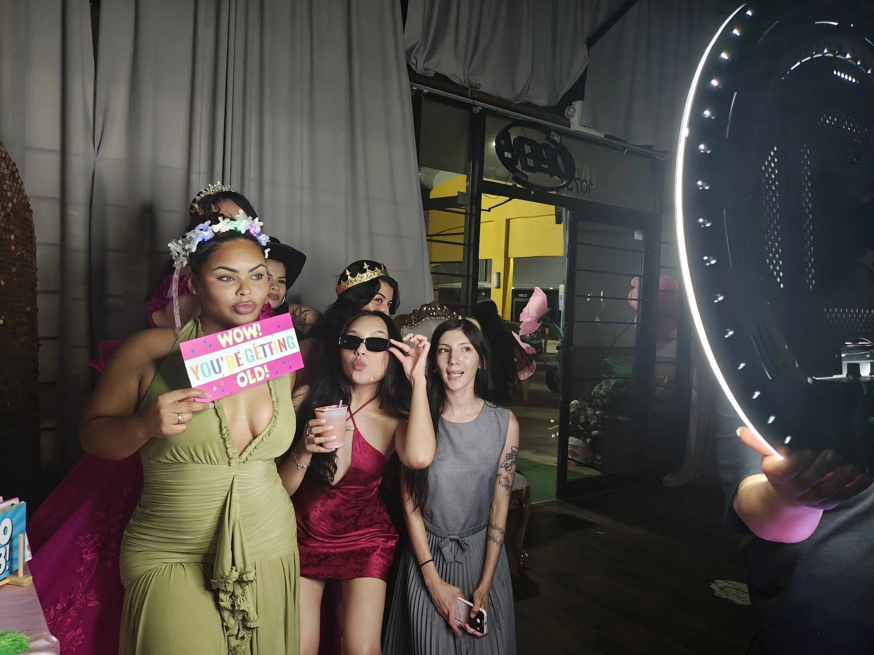 Group of five women posing for a photo in a studio with a gray curtain backdrop, some wearing crowns and costumes, one holding a sign that reads "WOW! YOU'RE GETTING OLD!", another wearing sunglasses, holding a drink, with a ring light visible on the right side.
