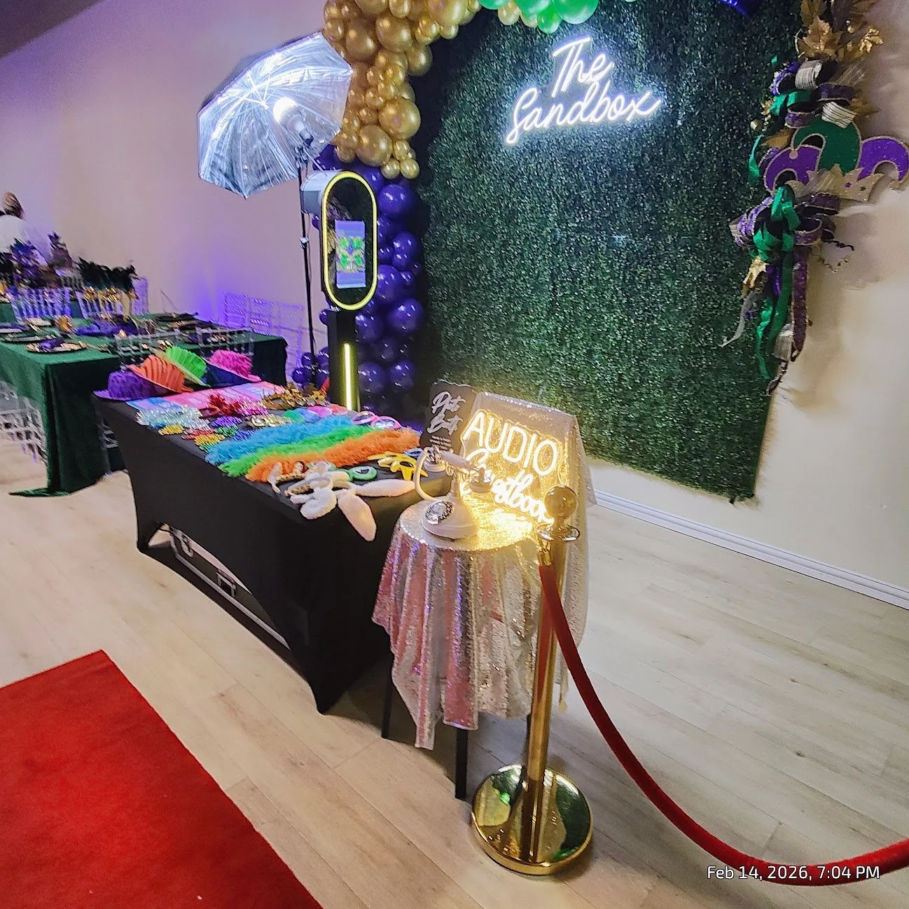 Decorative display at a party or event with a backdrop featuring green artificial foliage, neon sign reading 'The Sandbox,' and a table with various colorful accessories including feather boas, hats, accessories, and plush items. There is a small tab