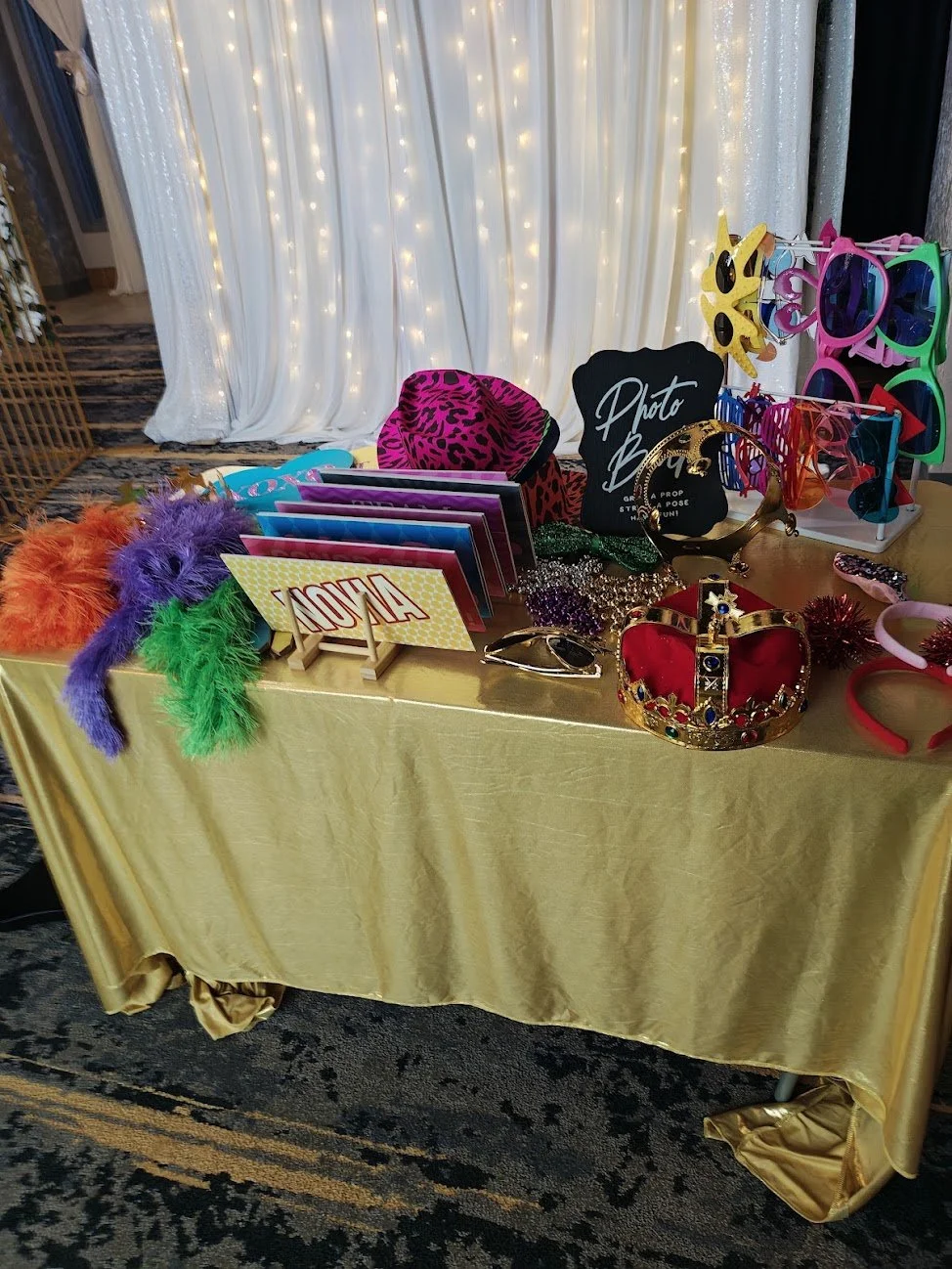 Photo booth prop table with colorful feather boas, sunglasses, a crown, hats, and a photo booth sign, set against a backdrop of white curtains with string lights.