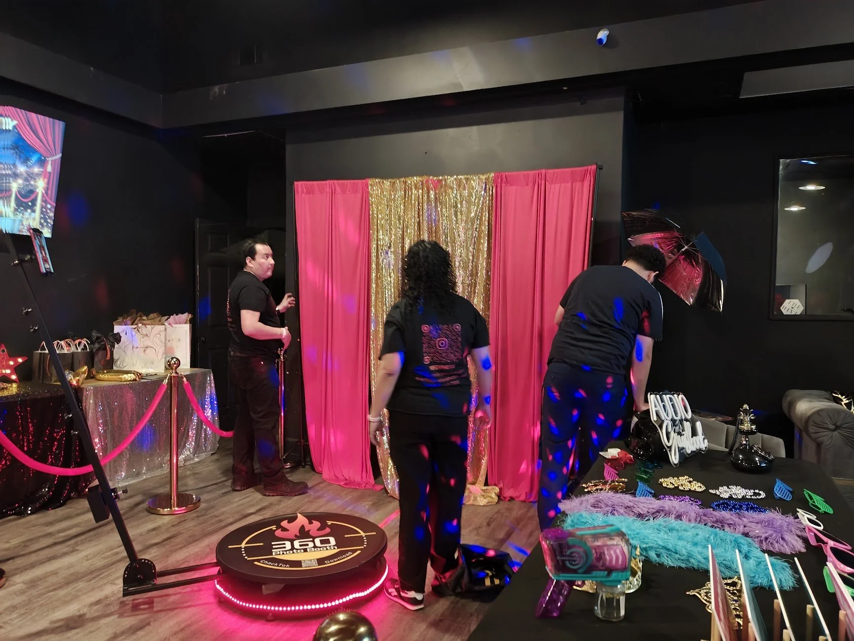 People at a celebration or party with a photo booth, props, and decorations, including pink curtains with gold accents, a table with gift bags, and various accessories like feather boas, glasses, and jewelry.