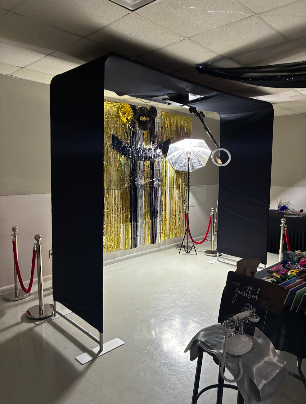 Photo of a photo booth setup with a black frame, gold and black decorations, a 'Happy 60th' banner, and an umbrella with lights.