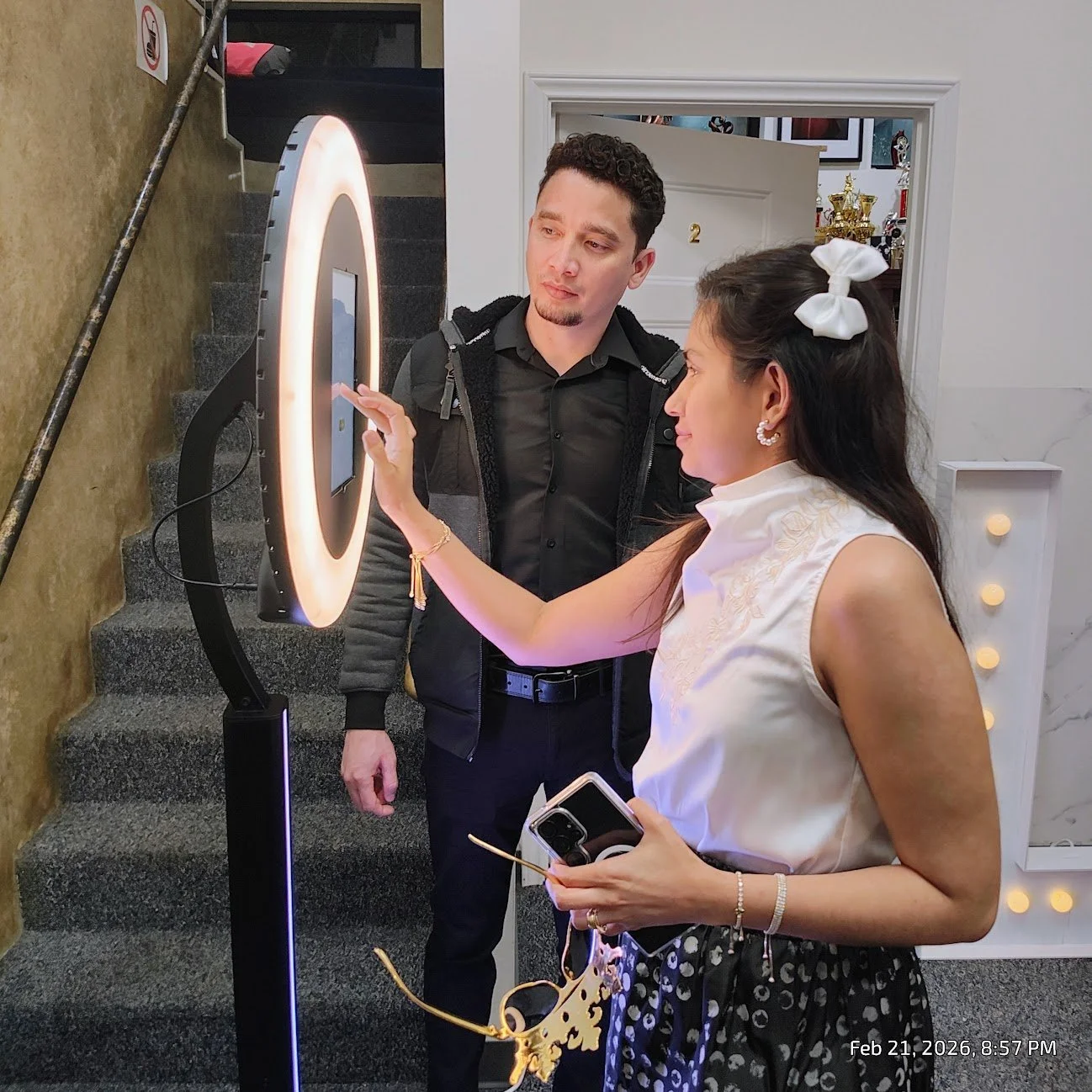 A woman is interacting with a photo booth selfie ring light station while holding a smartphone and glasses, and a man stands nearby observing. They are in an indoor space with stairs and decorated walls in the background.