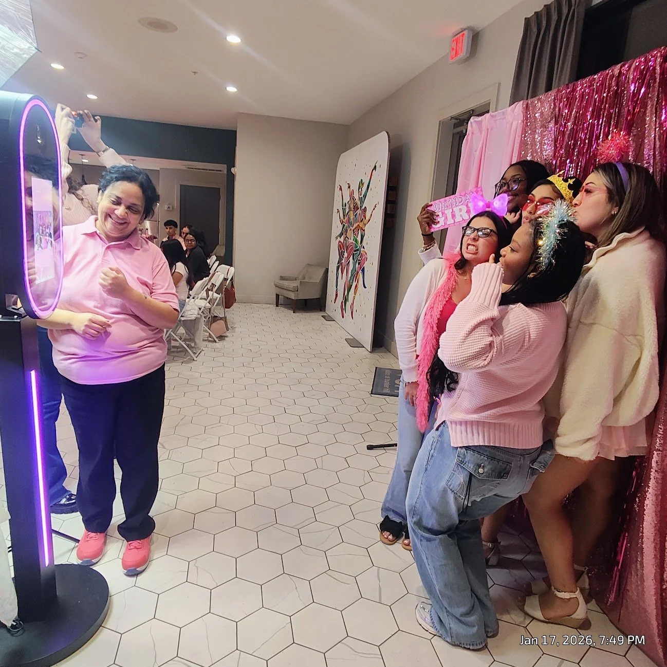 A group of women celebrating a birthday party, posing for a photo with a woman using a photo booth. The women are dressed in colorful clothes and accessories, some wearing headbands and scarves, and the background features a pink curtain and a pink '