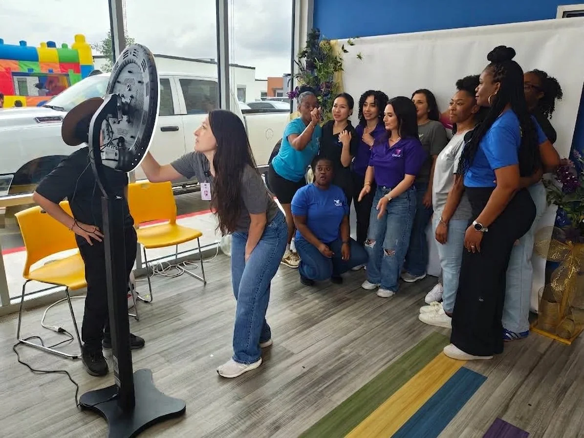 A group of women and girls posing for a photo while a woman adjusts a circular machine display, inside a building with large windows and a colorful indoor bounce house visible outside.