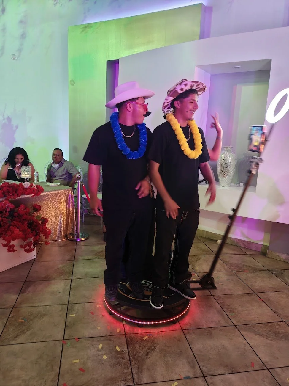 Two young men wearing large hats and colorful leis taking a selfie on a hoverboard at a celebration. People are sitting at a decorated table with flowers in the background.