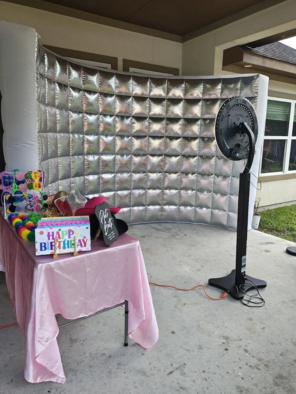 A photo booth setup with a silver quilted backdrop, a pink table with birthday props including a 'Happy Birthday' sign, colorful sunglasses, hats, and a chalkboard sign.